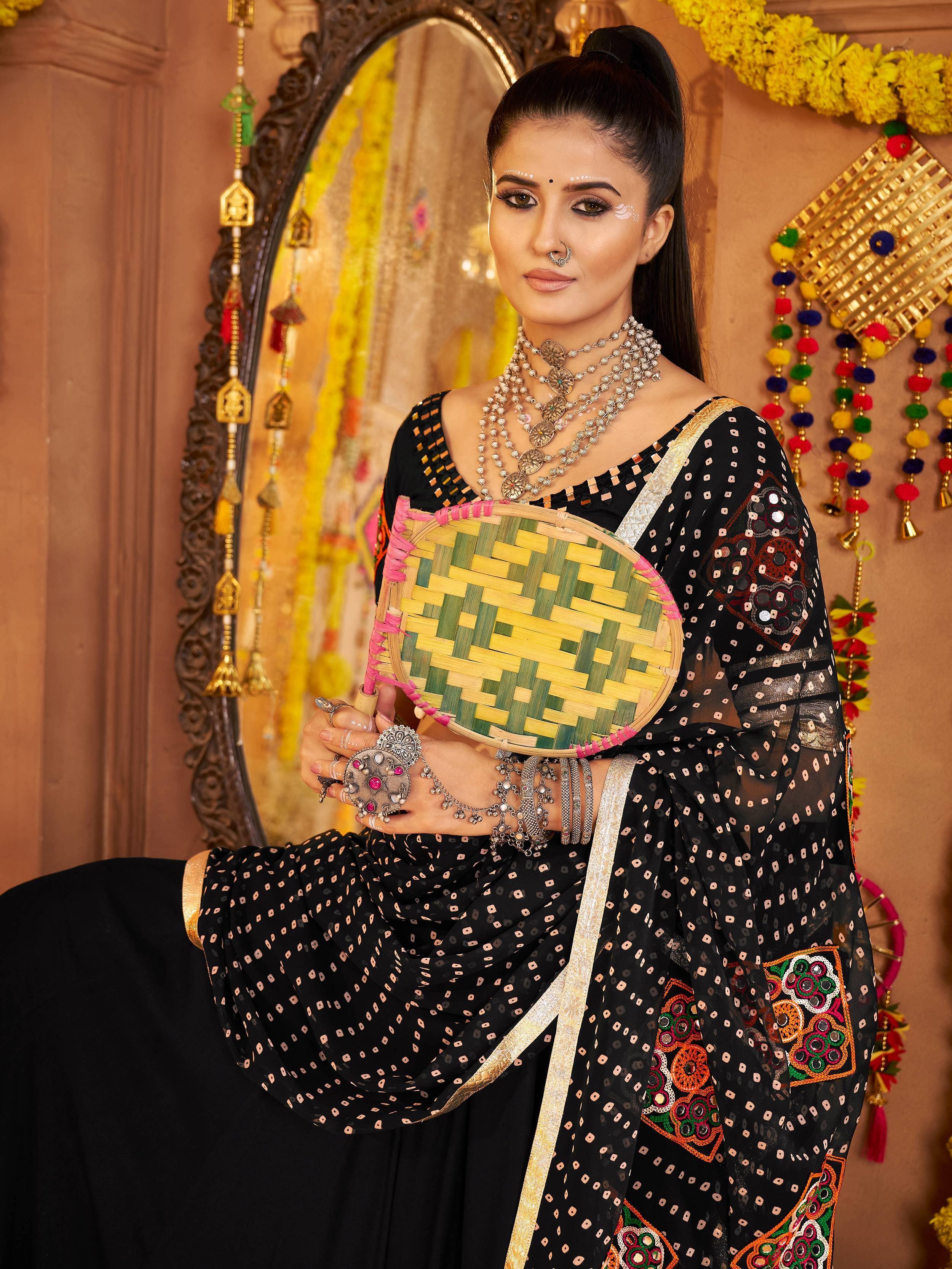 Woman in black embroidered ethnic saree with mirror work, holding woven fan, festive Indian decor