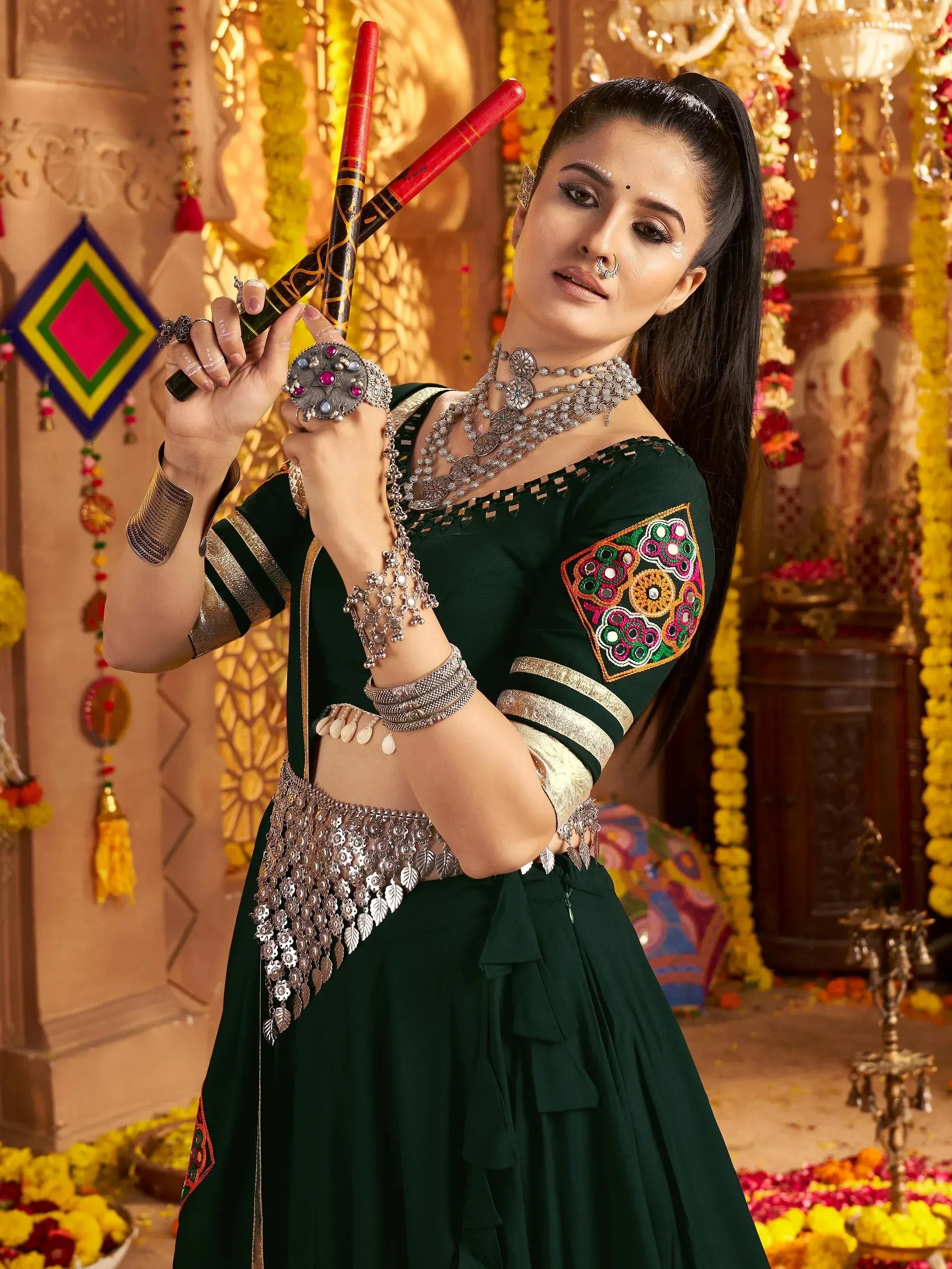 Woman in green embroidered lehenga choli with traditional jewelry and dandiya sticks for festive Indian wear