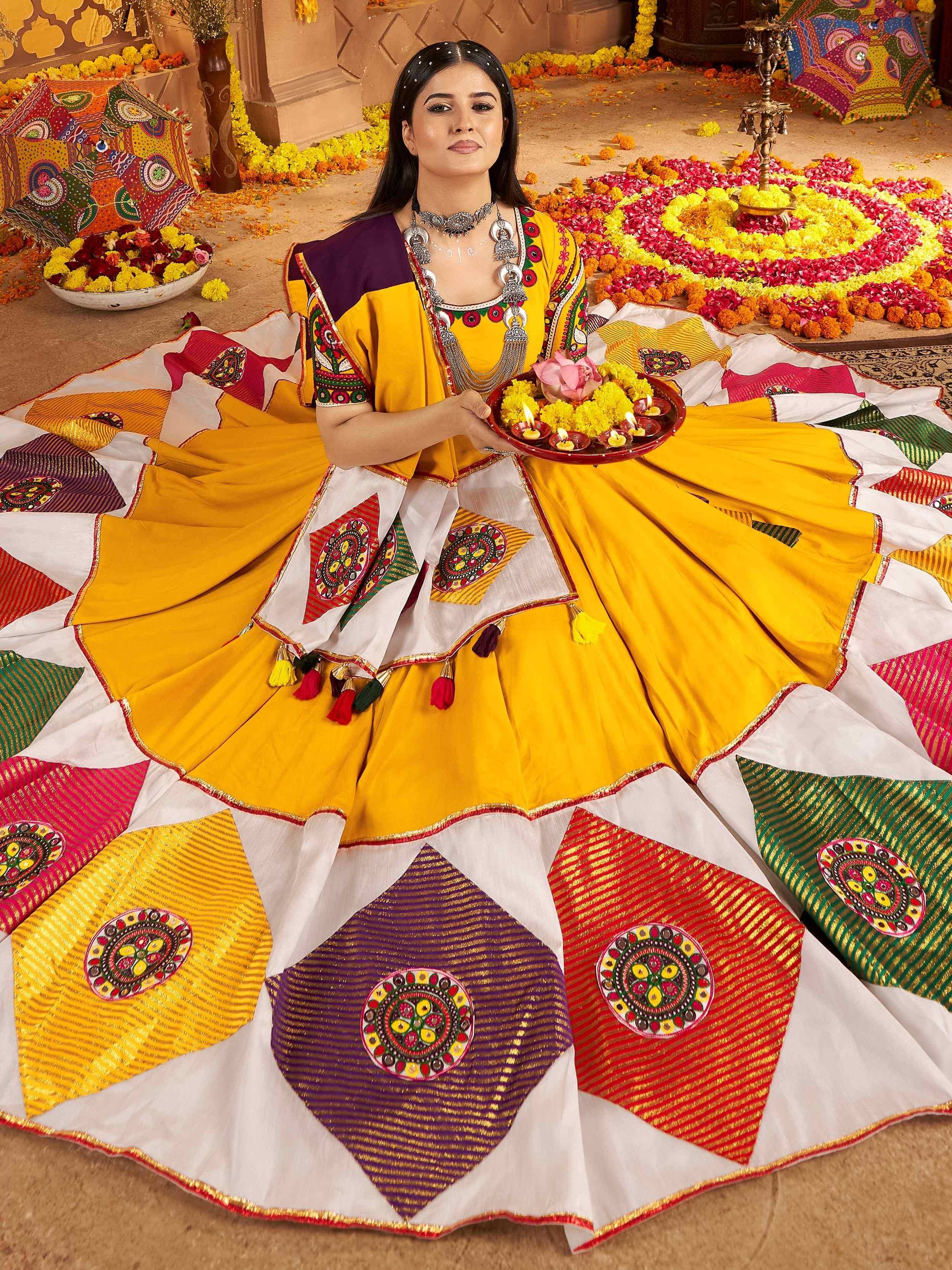 Woman in vibrant yellow and multicolor embroidered ethnic gown with floral rangoli, holding diya tray for festive celebration