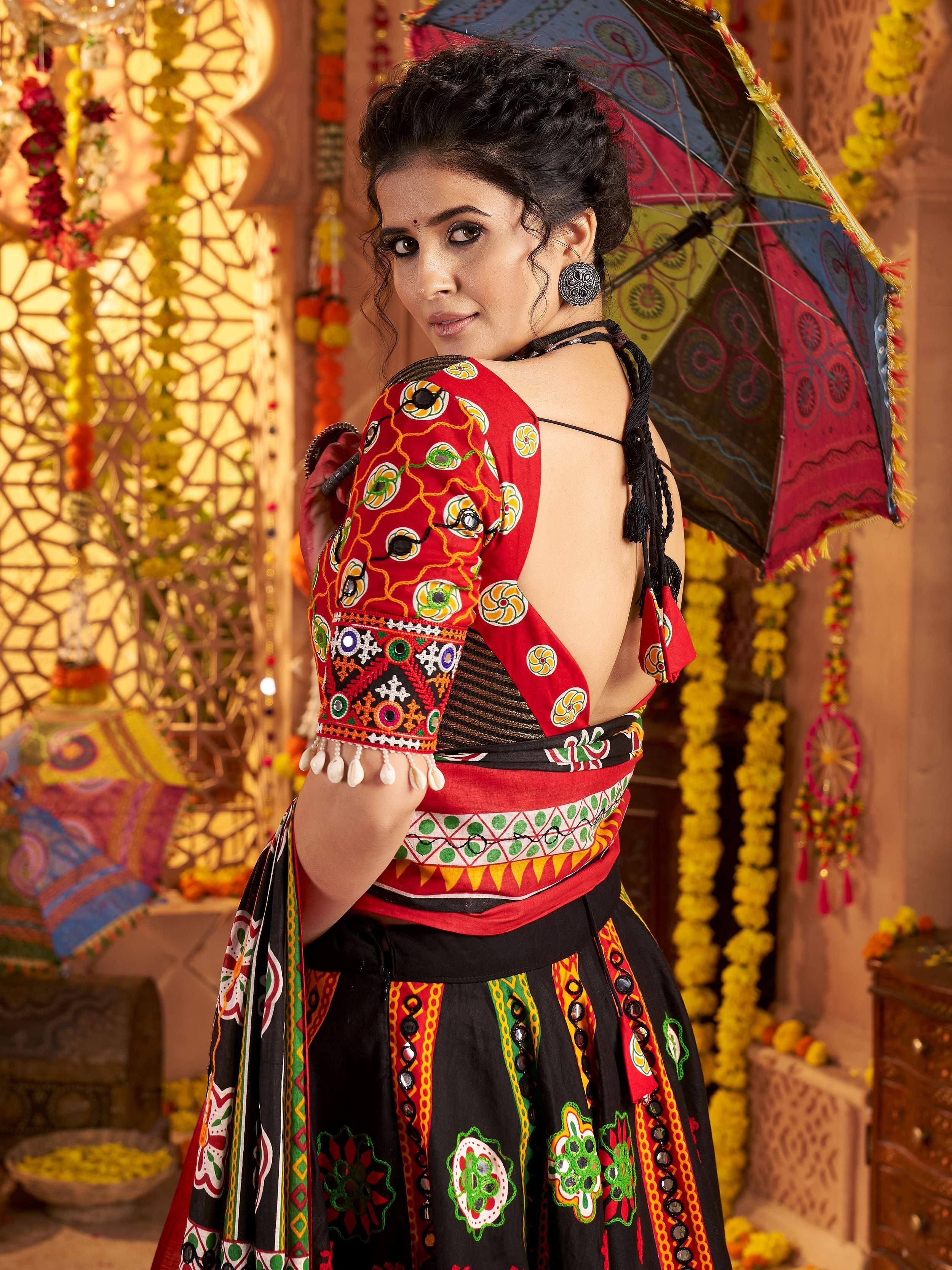 Woman in vibrant red and black embroidered ethnic wear with mirror work, holding decorative umbrella, festive Indian setting