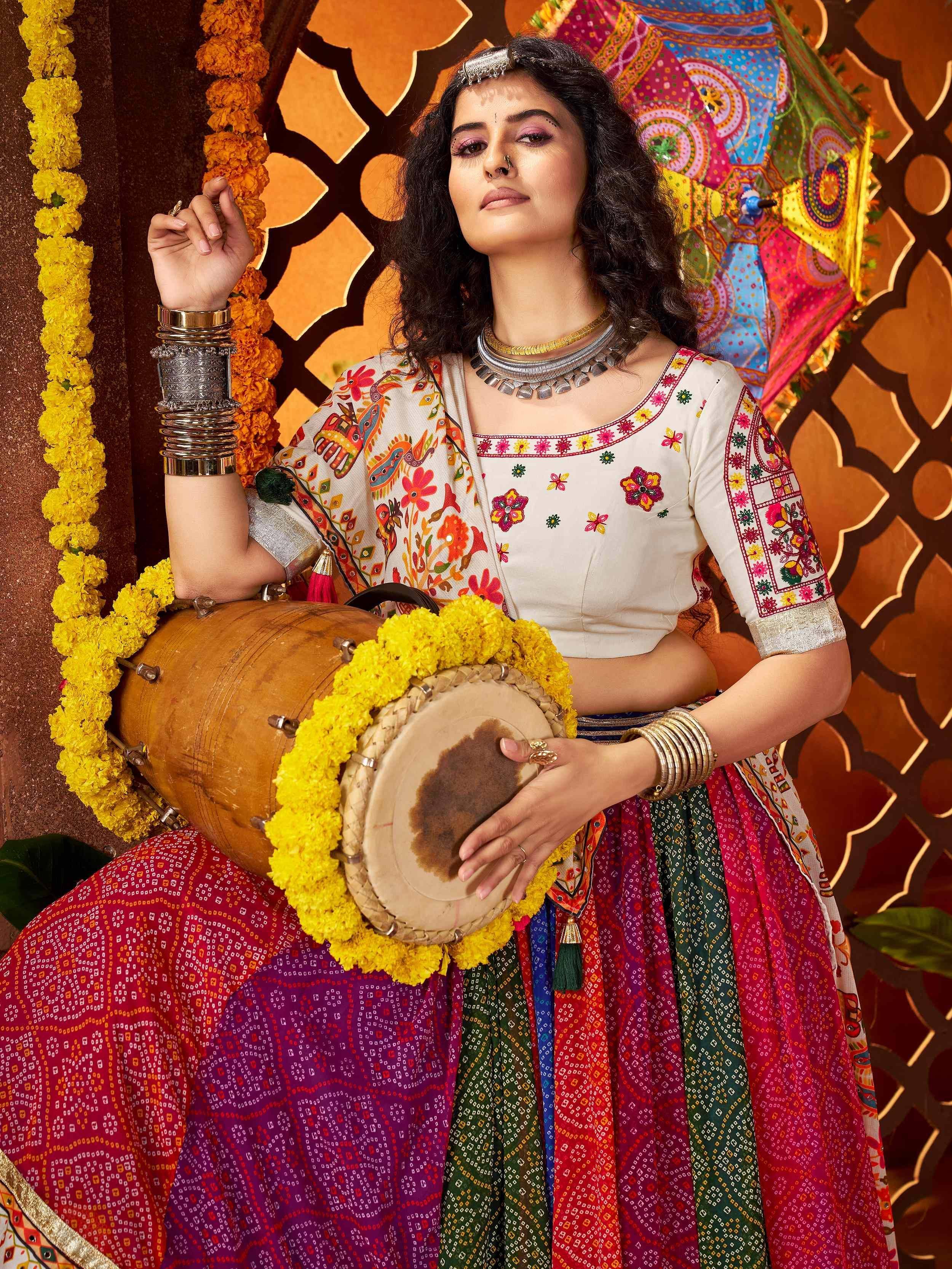 Woman in colorful Indian ethnic wear with floral embroidery, holding traditional drums, adorned with marigold garlands and tribal jewelry.