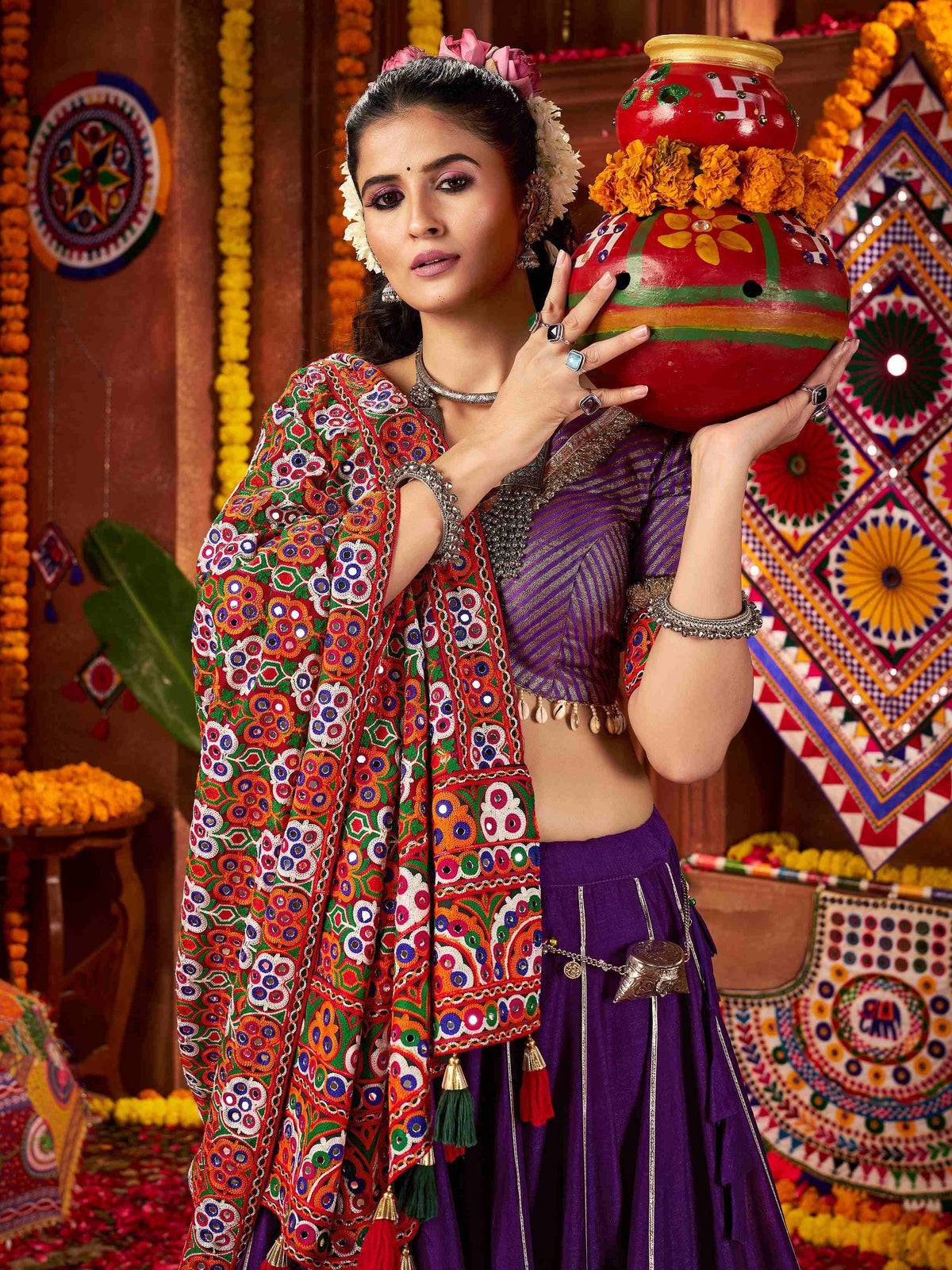 Woman in vibrant Indian ethnic wear holding decorated pot, floral headpiece, mirror work dupatta