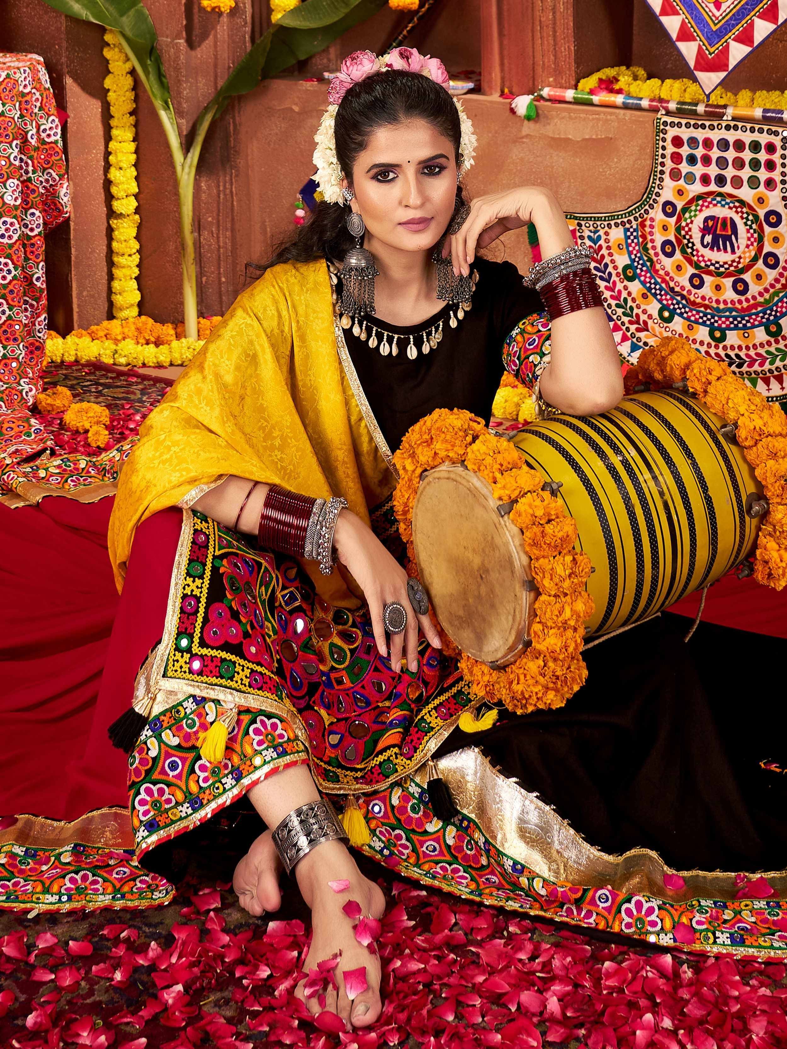 Woman in vibrant ethnic wear with mirror work, floral accessories, and traditional drum, surrounded by marigolds and rose petals.