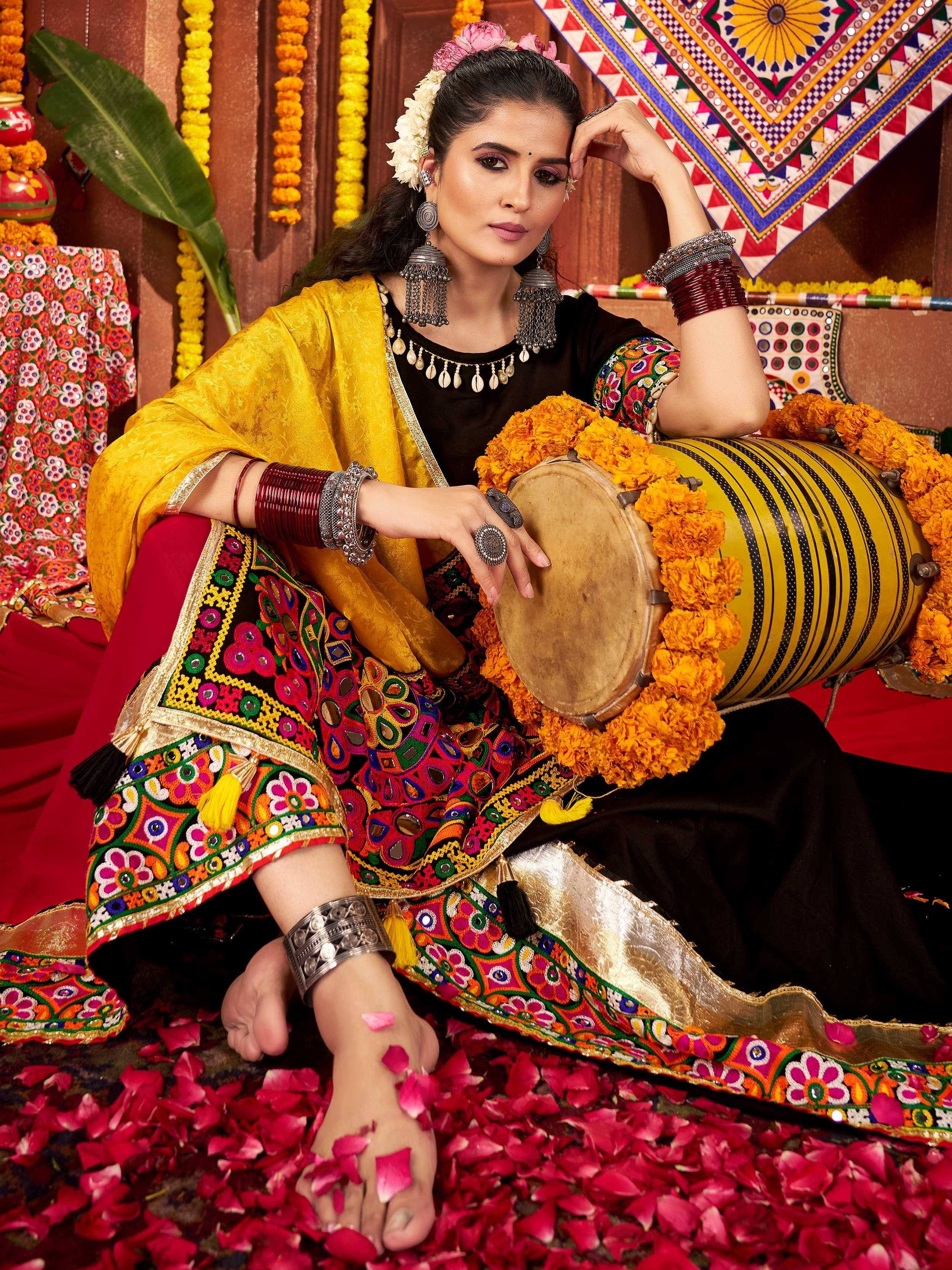 Woman in vibrant ethnic wear with mirror work, holding a decorated dhol, surrounded by marigold garlands and rose petals.