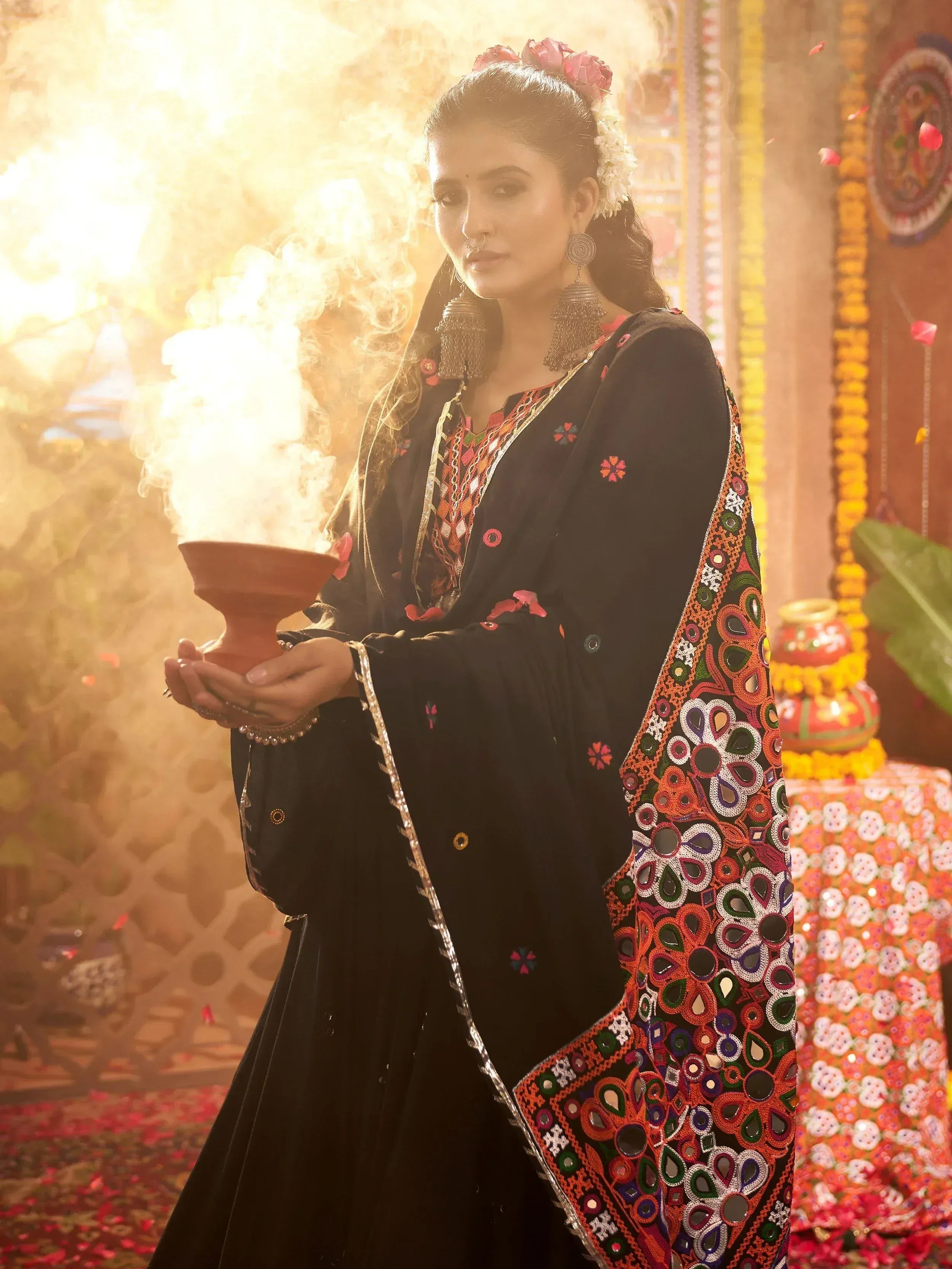 Woman in black embroidered ethnic gown holding smoking clay pot, festive Indian decor