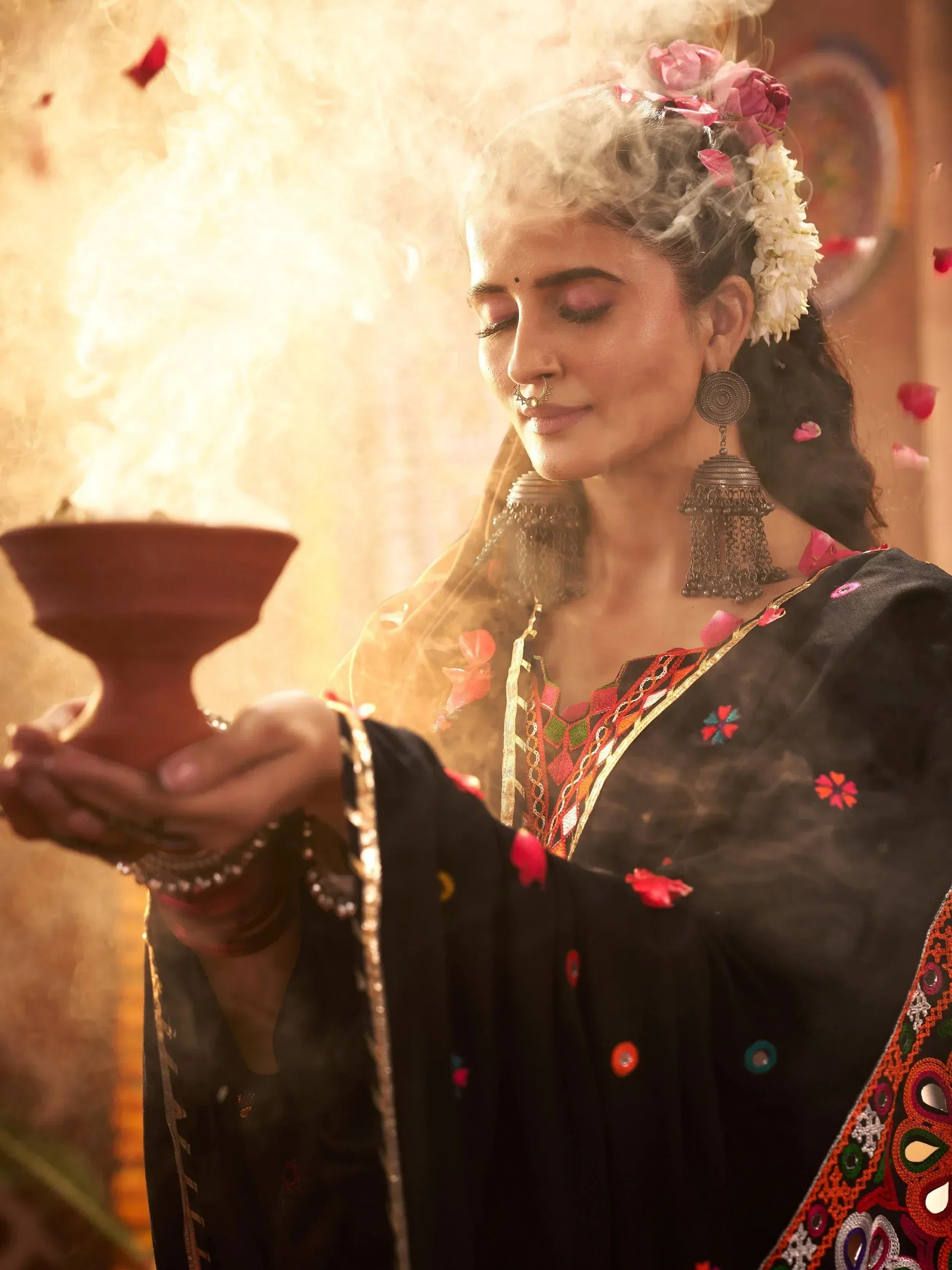 Woman in black embroidered ethnic gown holding incense bowl with floral hair adornment