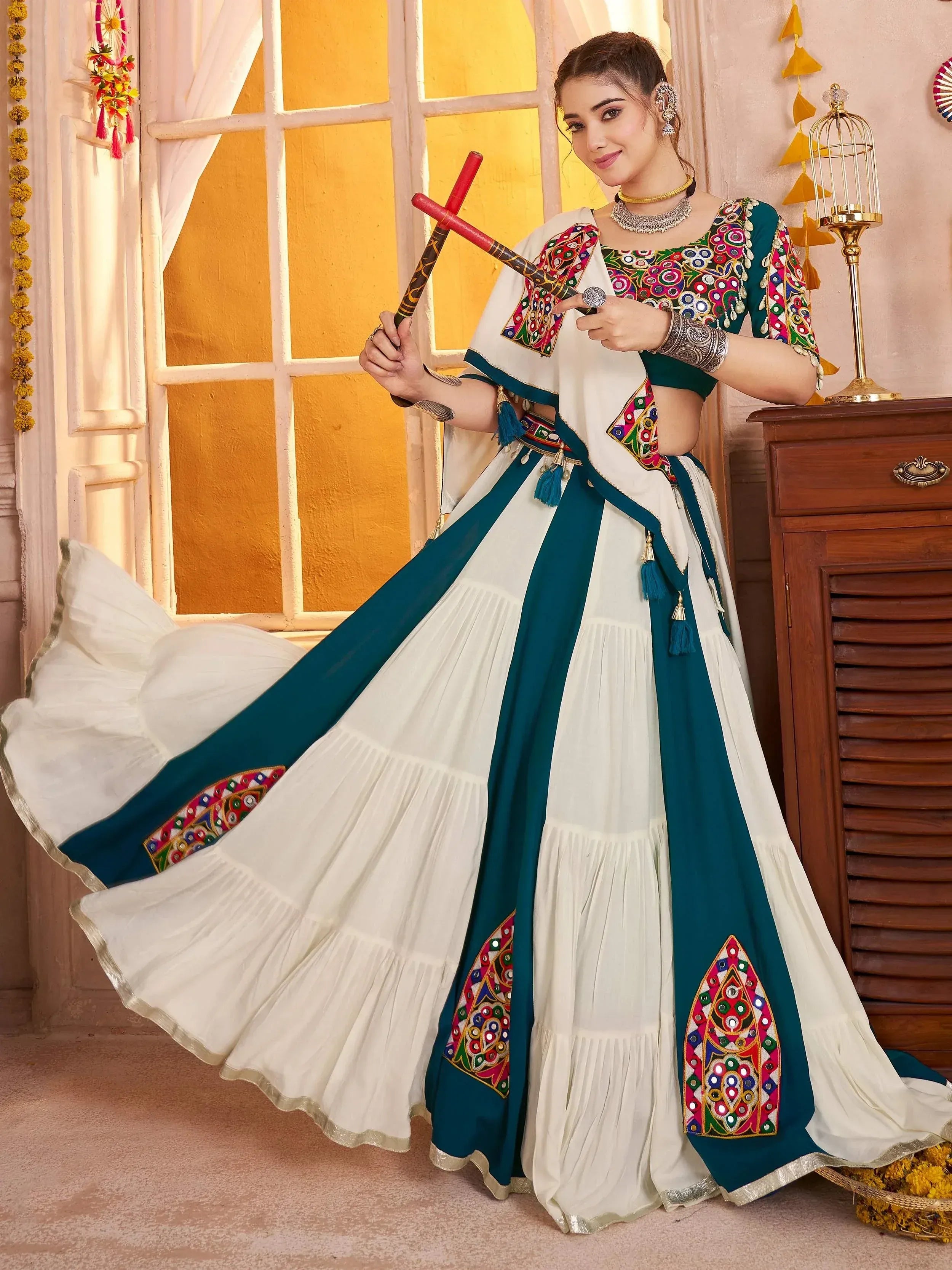 Woman in white and teal embroidered lehenga choli with mirror work, holding dandiya sticks for festive wear
