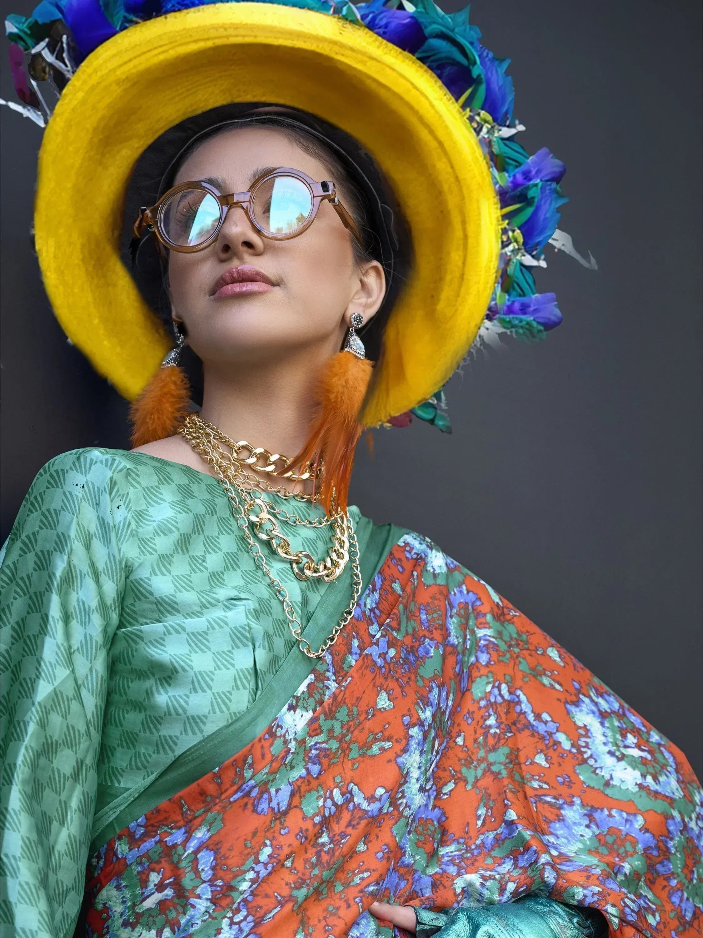 Woman in vibrant printed saree with yellow floral hat, gold chains, and round glasses