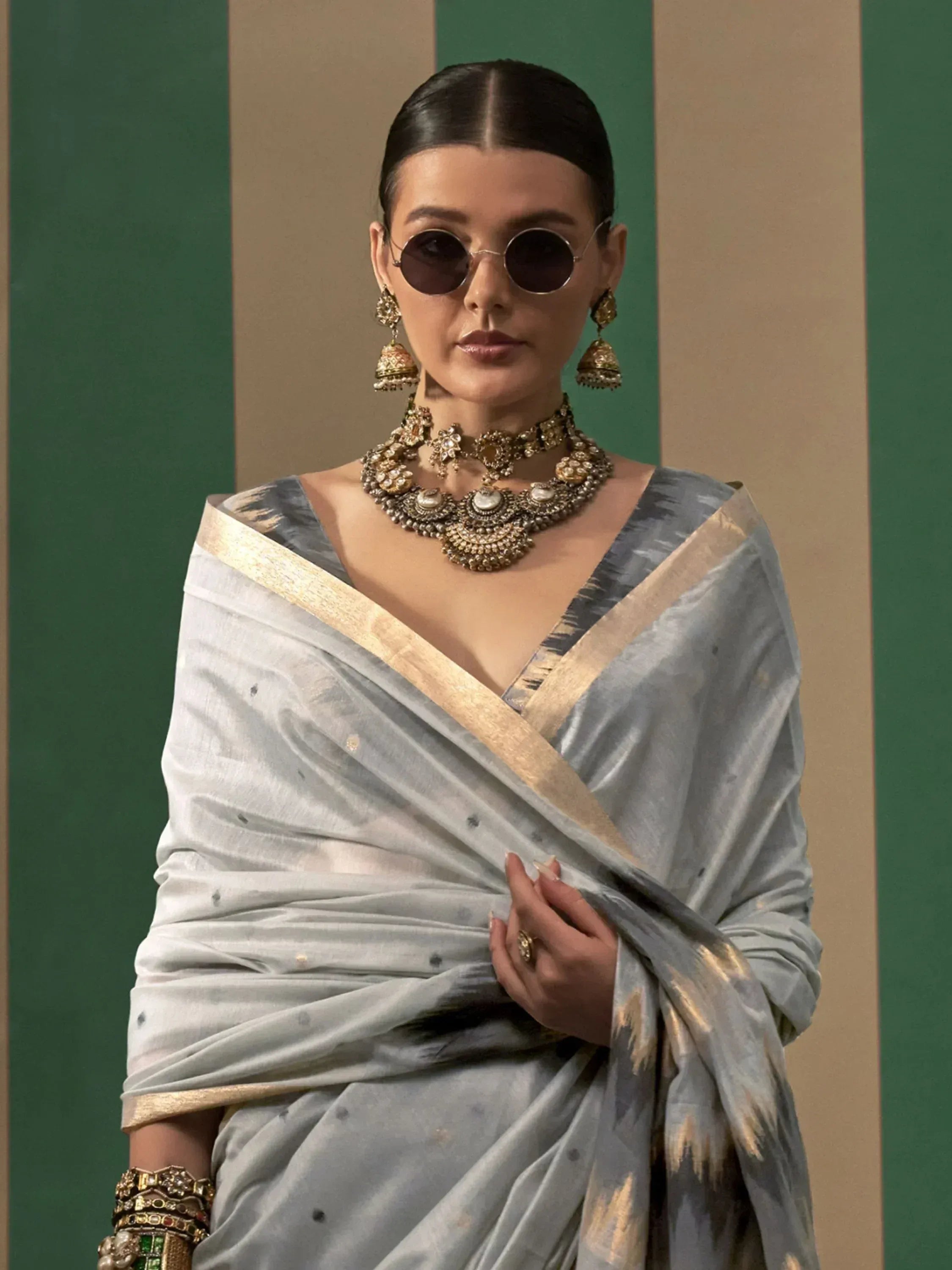 Woman in elegant grey saree with gold border, traditional jewelry, and round sunglasses
