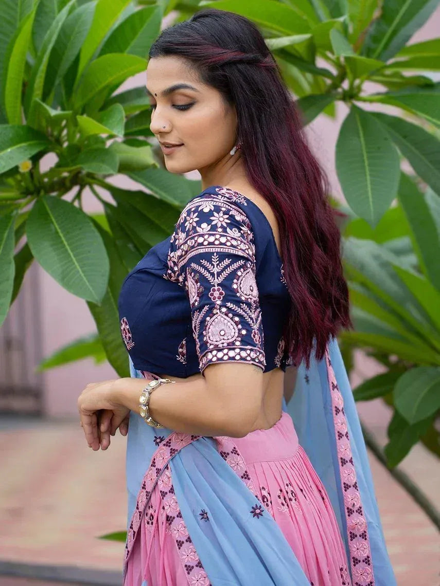 Woman in blue and pink embroidered lehenga choli with floral patterns, standing outdoors near green foliage