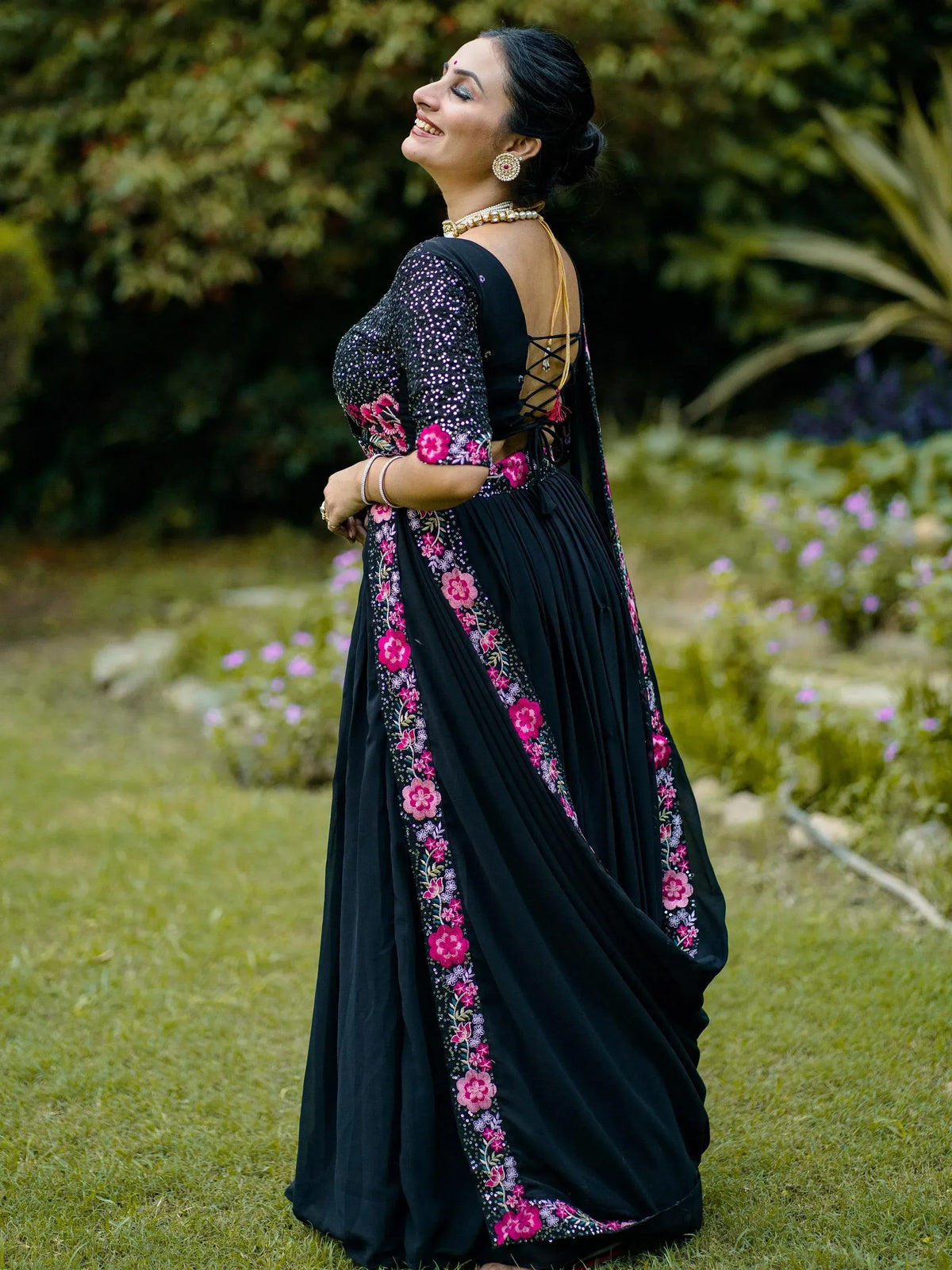 Black ethnic gown with pink floral embroidery, sequin sleeves, and lace-up back, worn in garden setting
