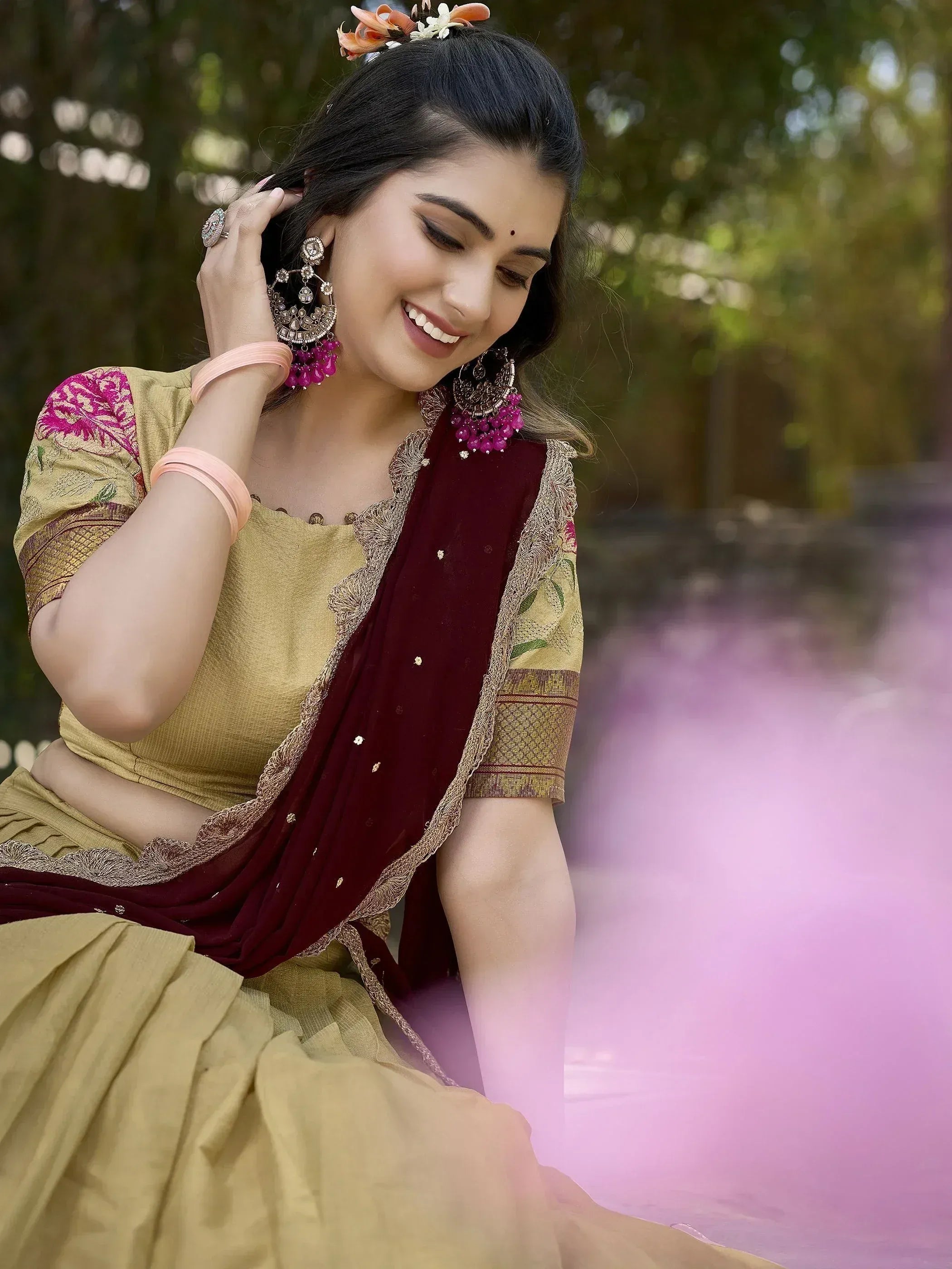 Woman in mustard yellow lehenga choli with maroon dupatta, floral hair accessories, and pink jhumka earrings.