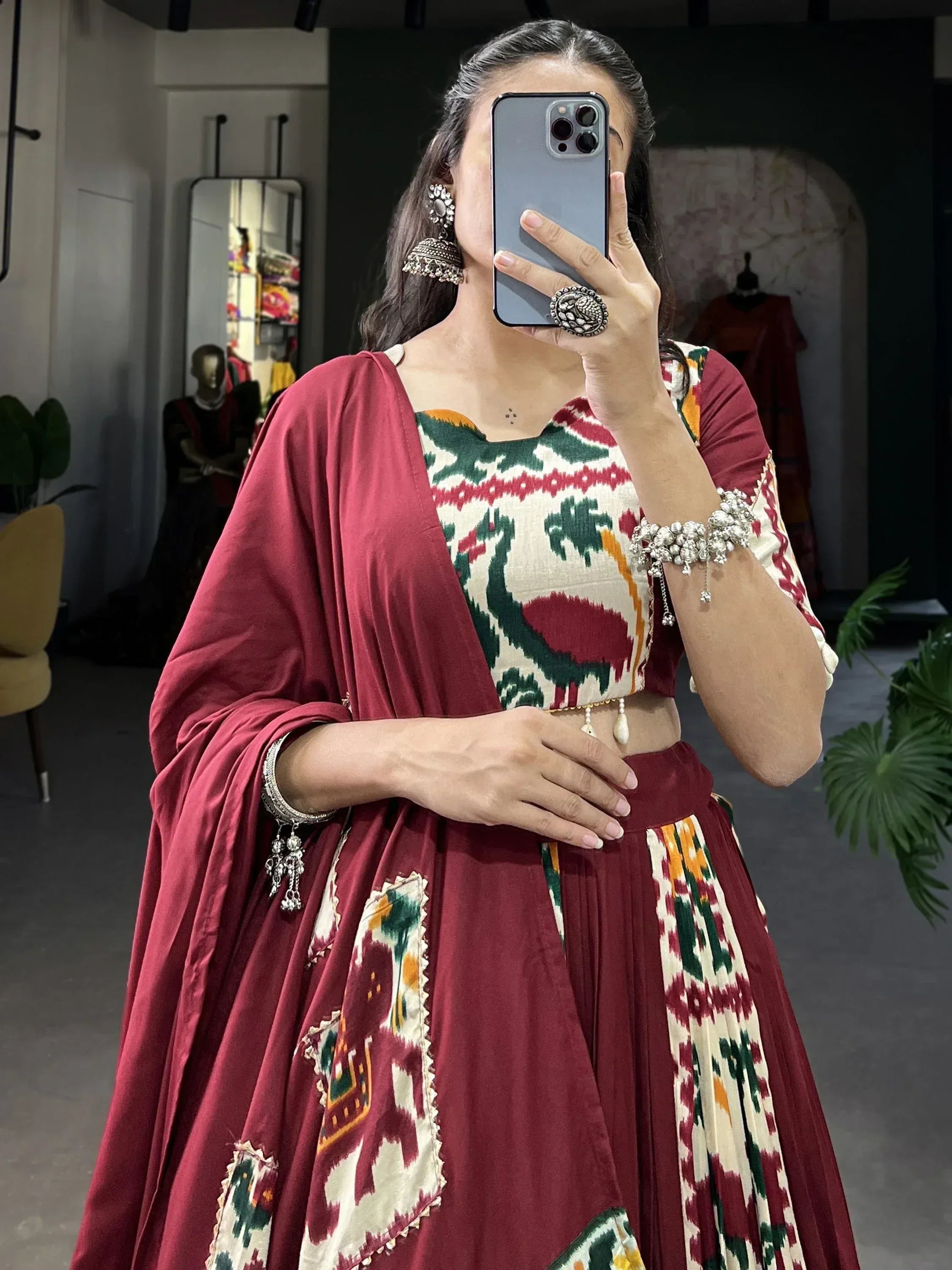 Woman in maroon Ikat lehenga choli with ethnic jewelry, mirror selfie in boutique
