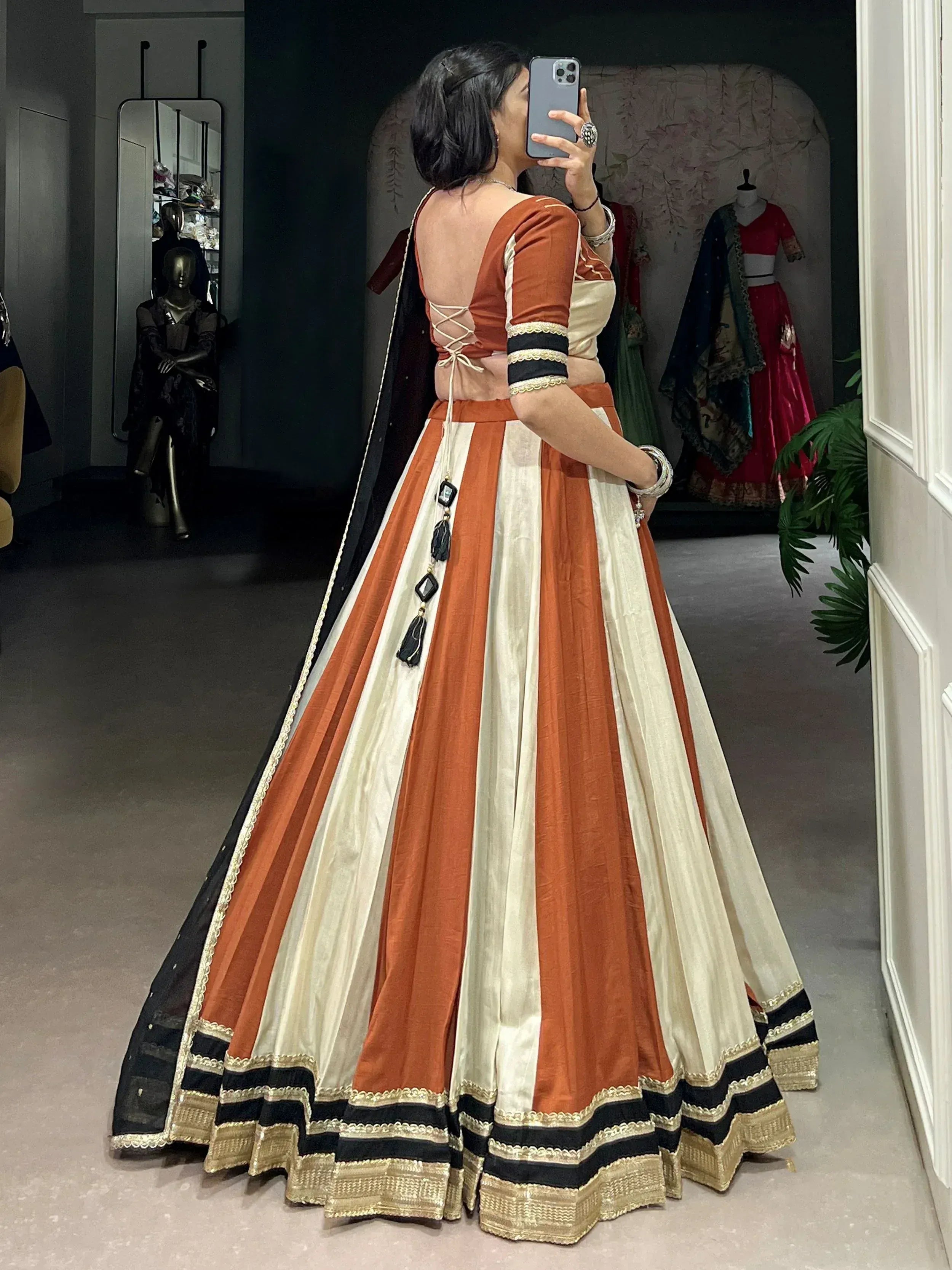 Woman in orange and cream striped lehenga choli with back lace-up detail, taking mirror selfie in boutique