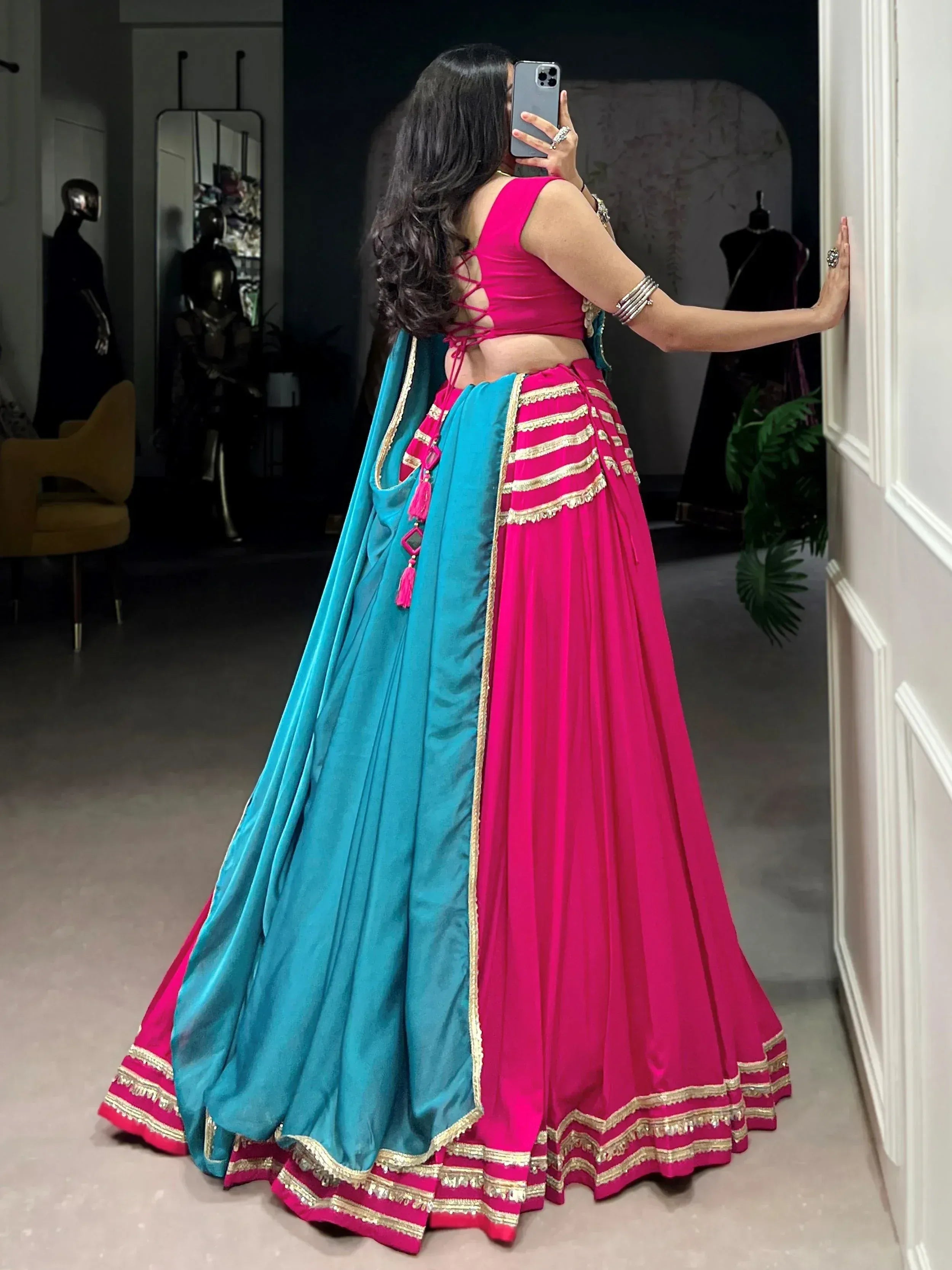 Woman in vibrant pink and teal lehenga choli with gold trim, taking mirror selfie in boutique