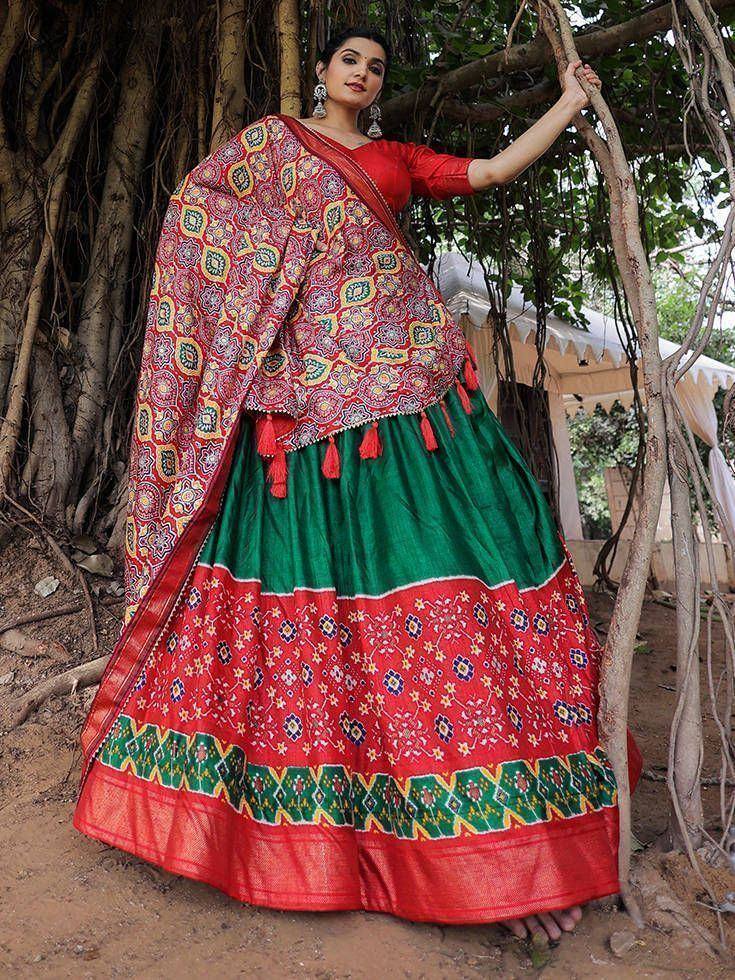 Woman in vibrant red and green ethnic lehenga choli with printed dupatta, standing near tree roots