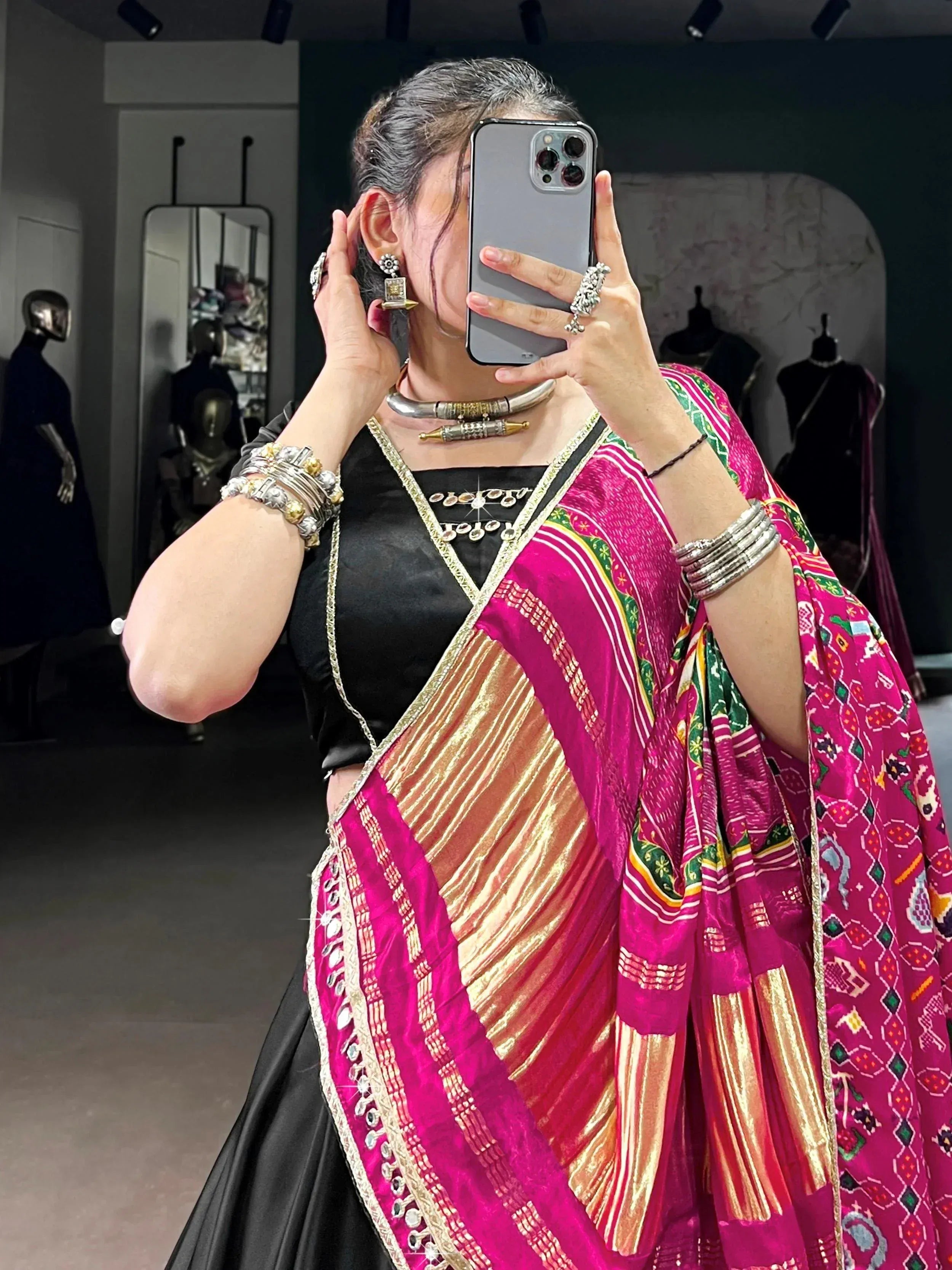Woman in vibrant pink and gold ethnic saree with mirror work, paired with black blouse and traditional jewelry, taking mirror selfie.