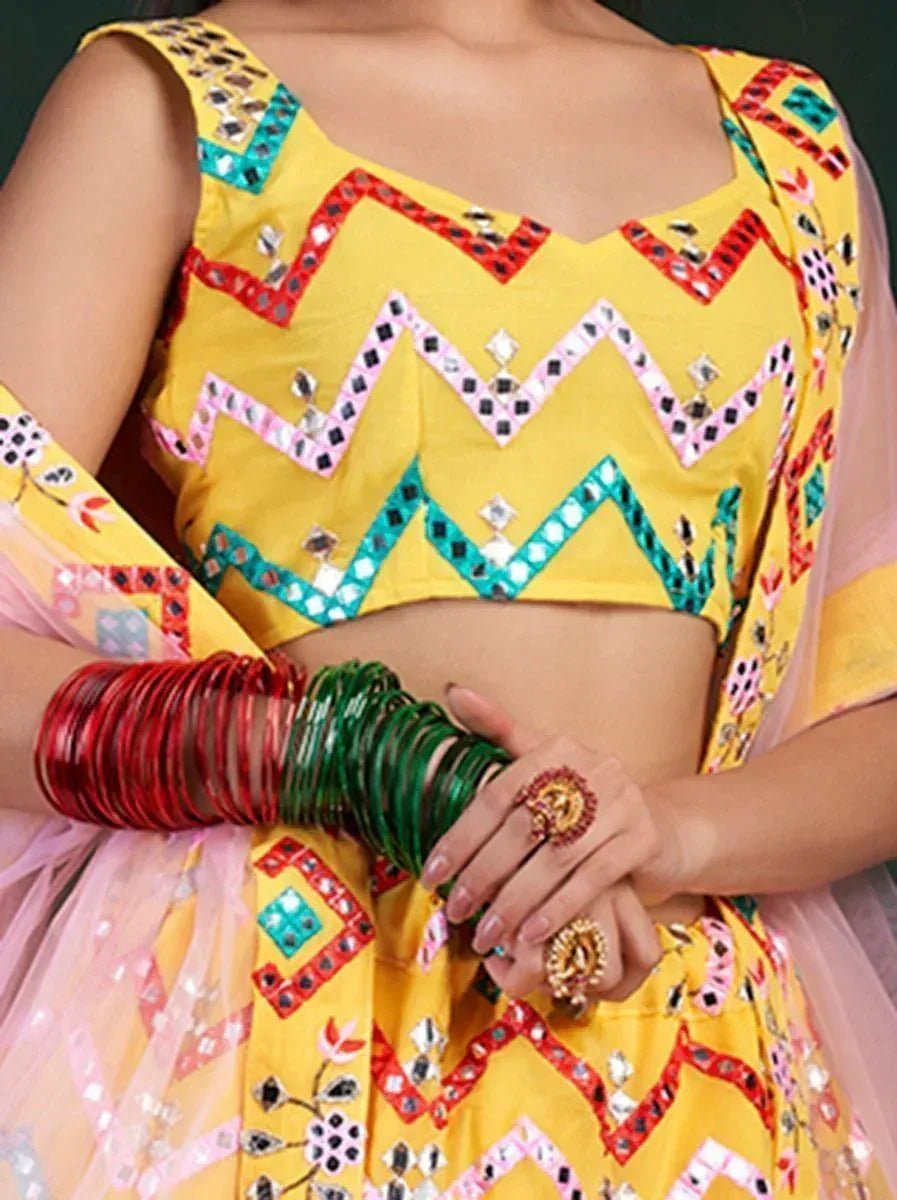 Yellow mirror-work lehenga choli with colorful zigzag embroidery, red and green bangles, and statement rings