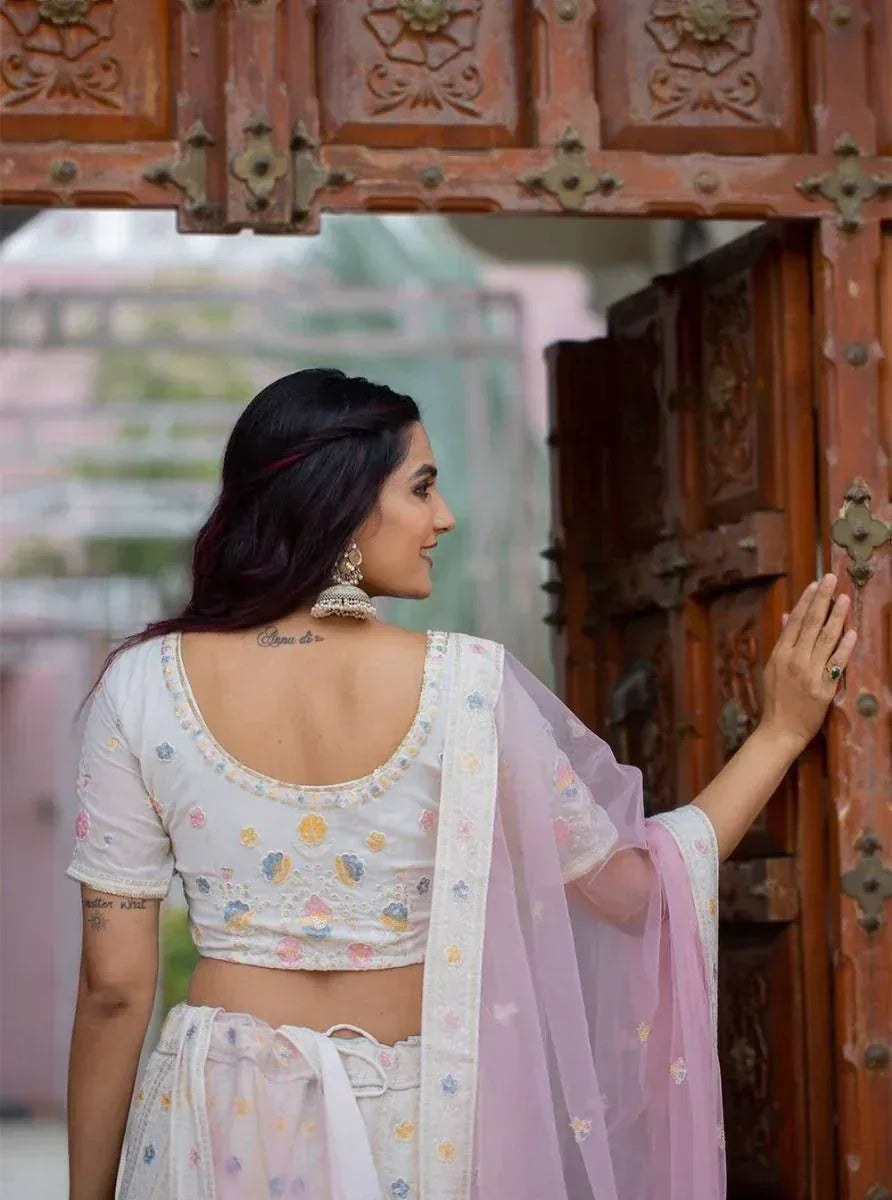 Woman in white floral embroidered lehenga choli with pink dupatta, standing by ornate wooden door