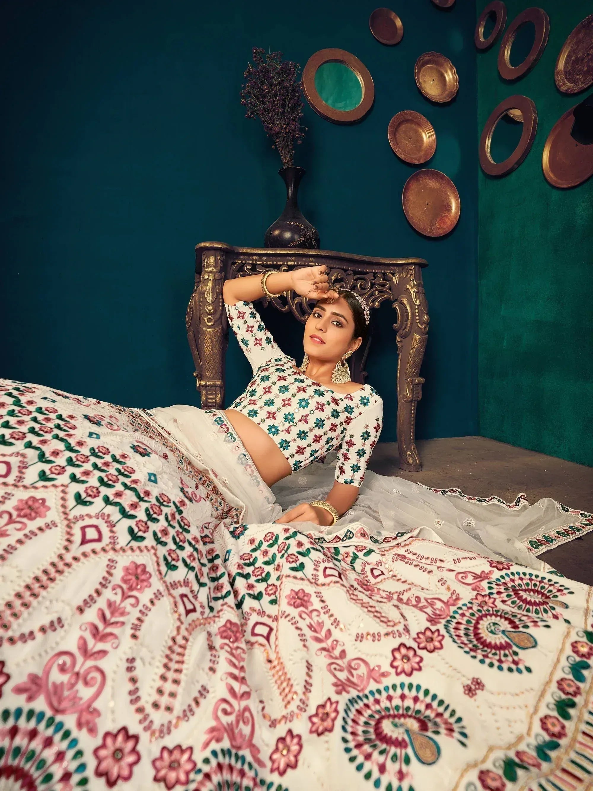 Woman in white floral embroidered lehenga choli with pink and green motifs, posing elegantly against teal backdrop.