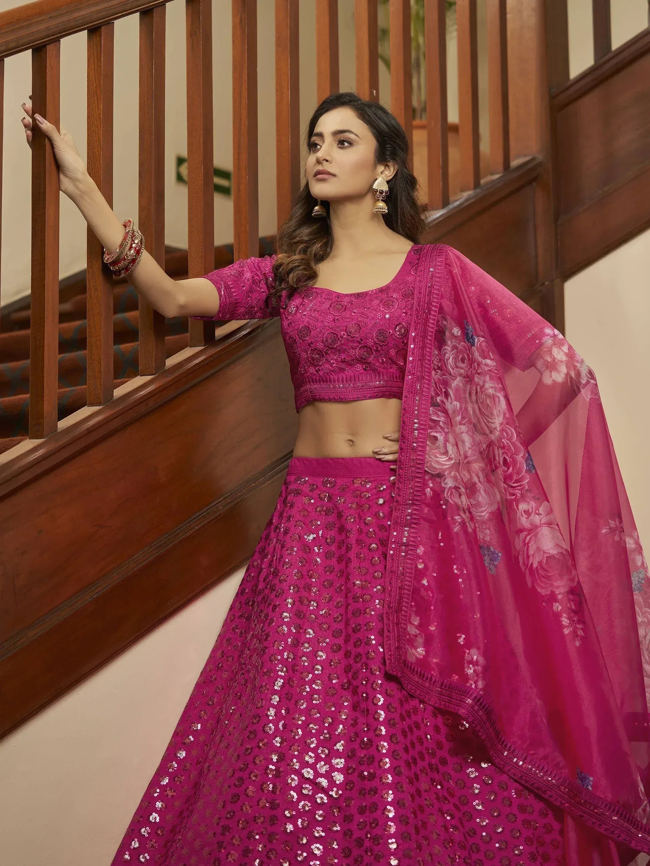 Woman in vibrant pink sequined lehenga choli with floral dupatta, posing by wooden staircase
