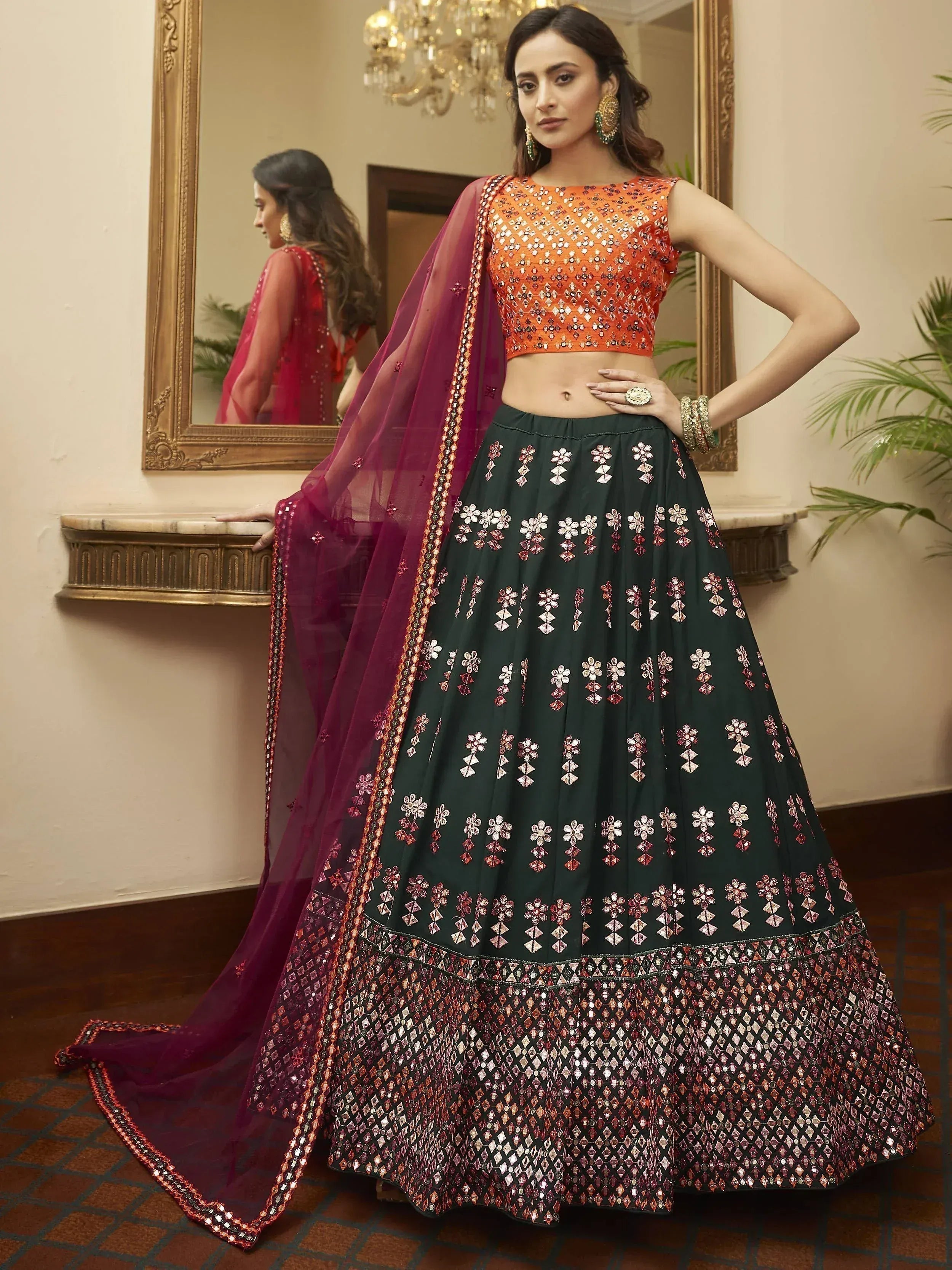 Designer lehenga choli with orange blouse, green skirt, and maroon dupatta, featuring mirror work and floral embroidery.