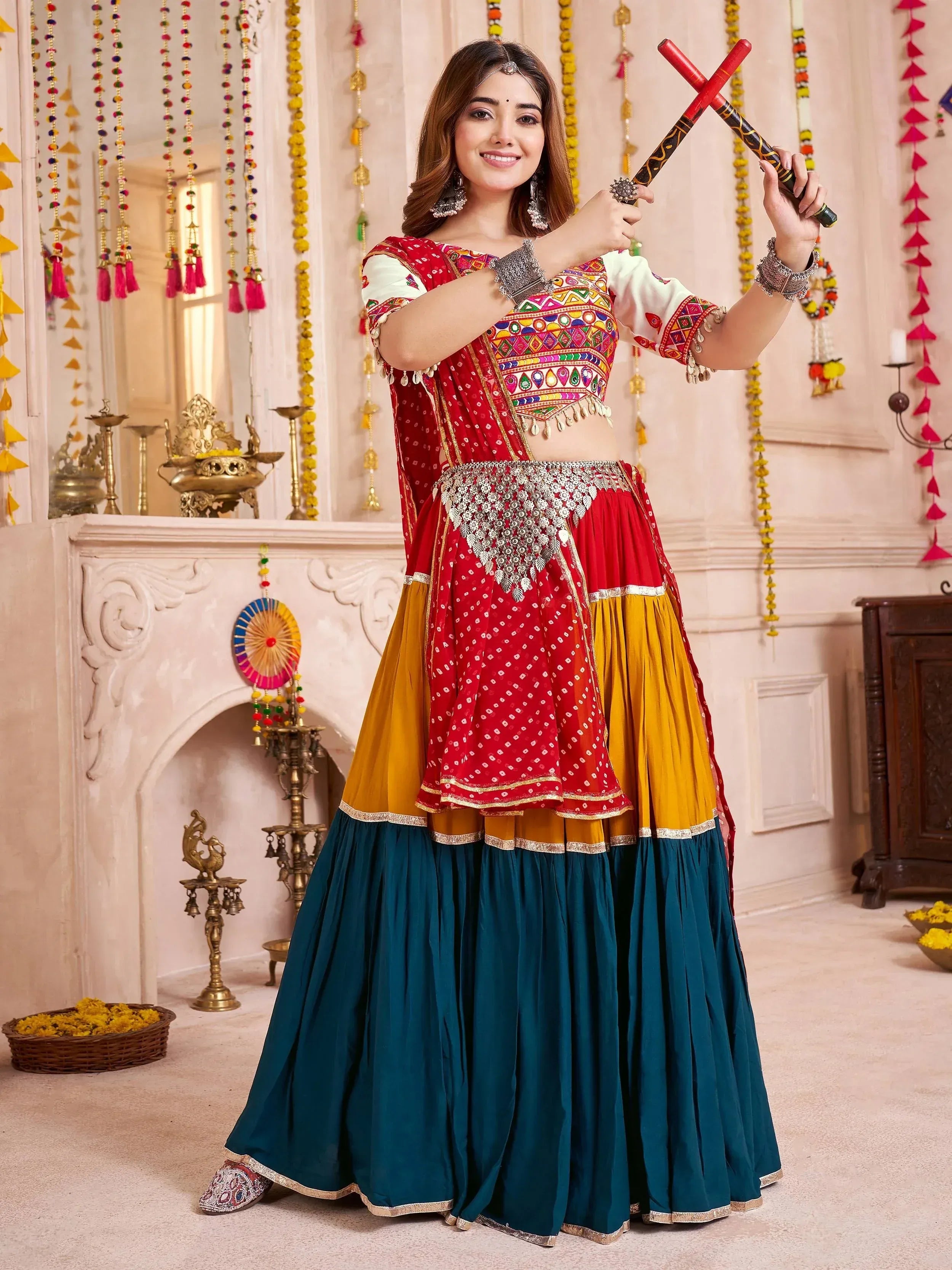 Woman in vibrant red, yellow, and teal Indian lehenga choli with mirror work, holding dandiya sticks for festive dance.