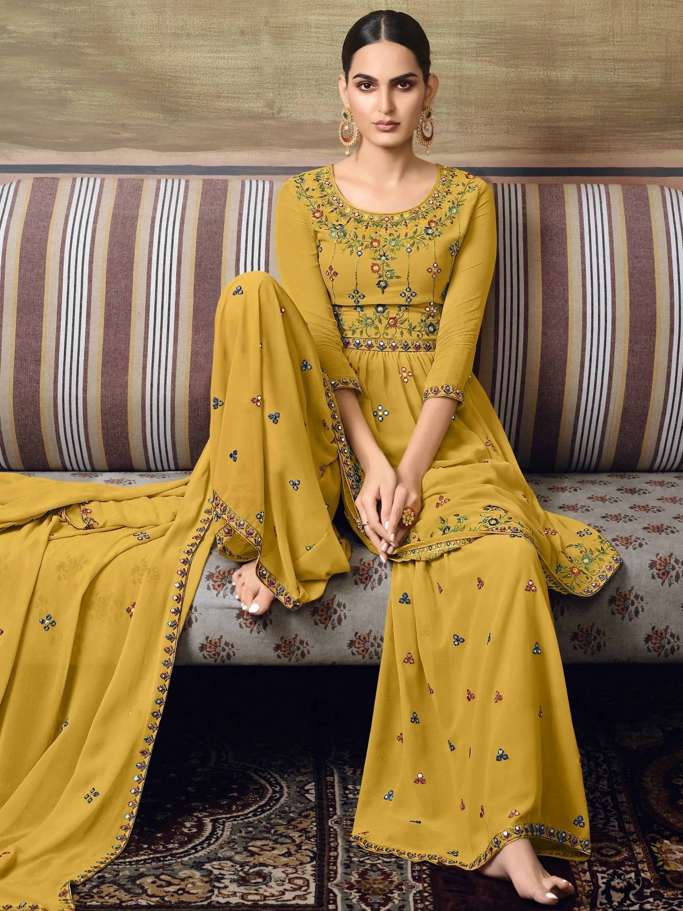 Woman in mustard yellow embroidered salwar kameez with dupatta, seated indoors
