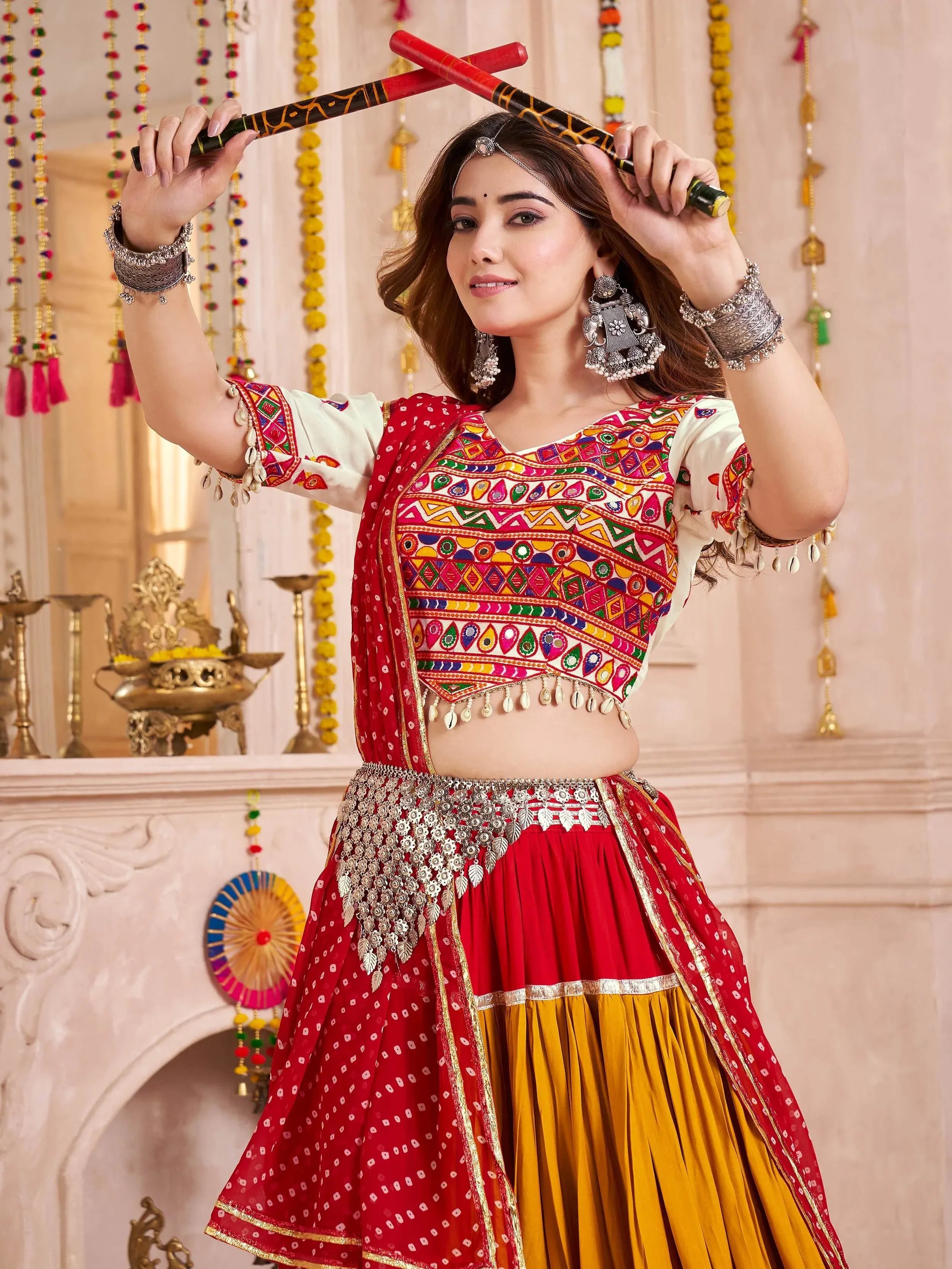 Woman in vibrant red and yellow ethnic lehenga choli with mirror work, holding dandiya sticks for festive dance