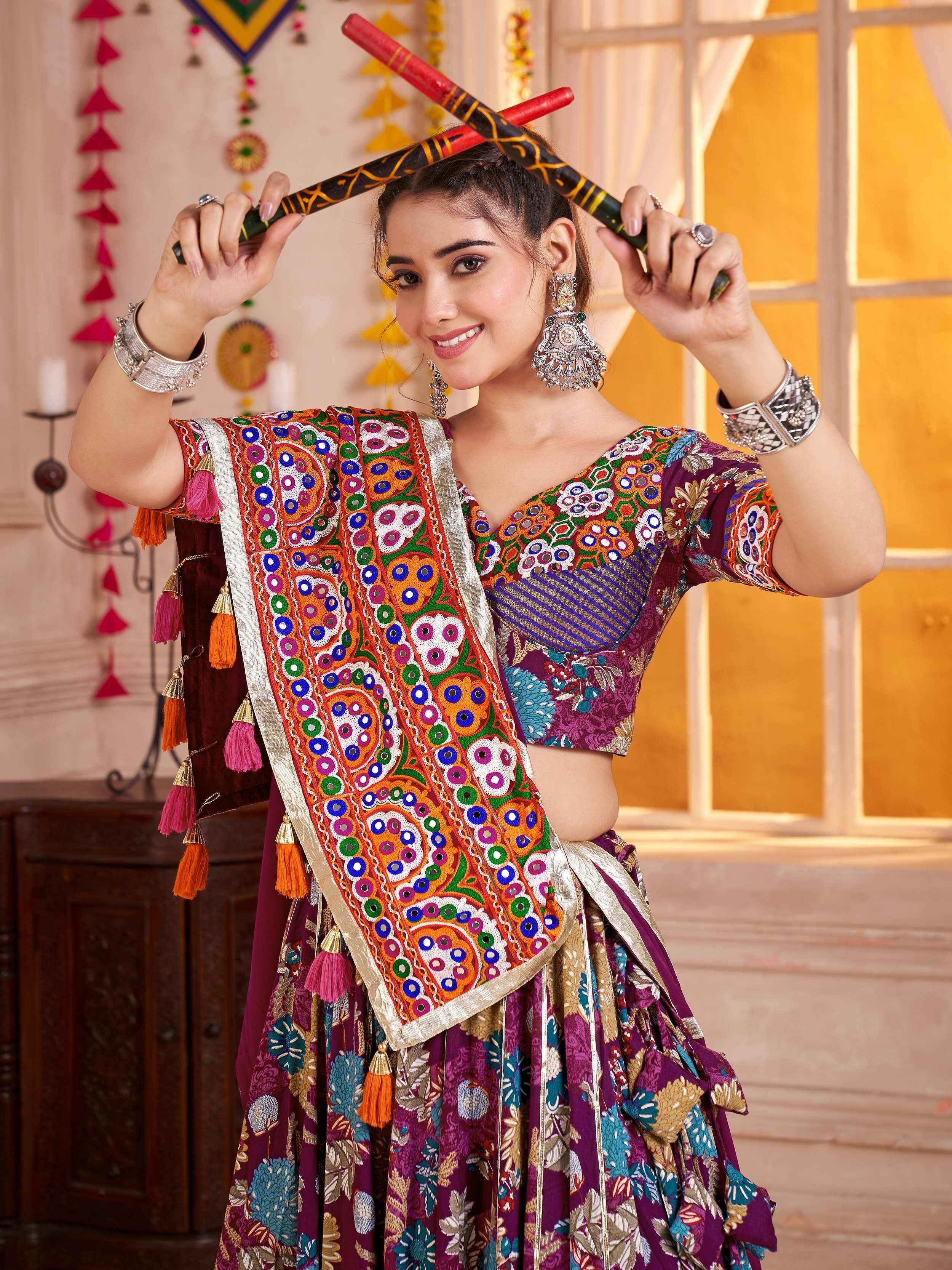 Woman in vibrant Indian ethnic lehenga choli with mirror work, floral prints, and tassels, holding decorative sticks.
