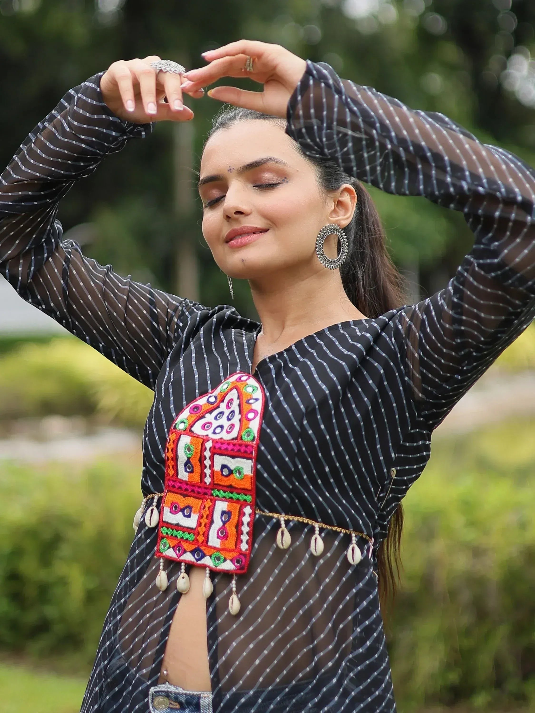 Woman in black striped ethnic top with colorful mirror work and shell embellishments, styled with statement earrings.