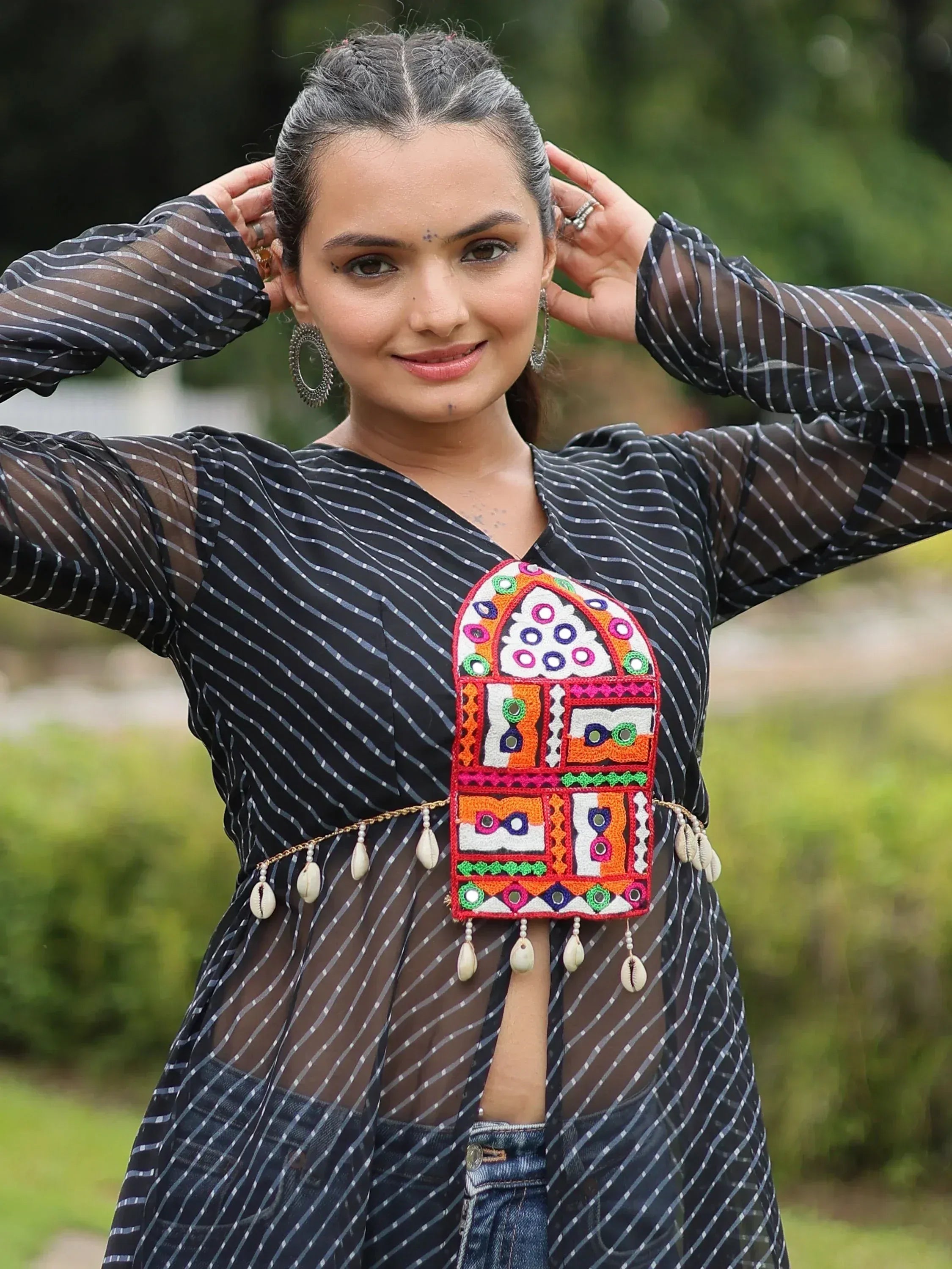 Woman in black striped sheer top with colorful embroidered ethnic belt and cowrie shells, outdoor setting