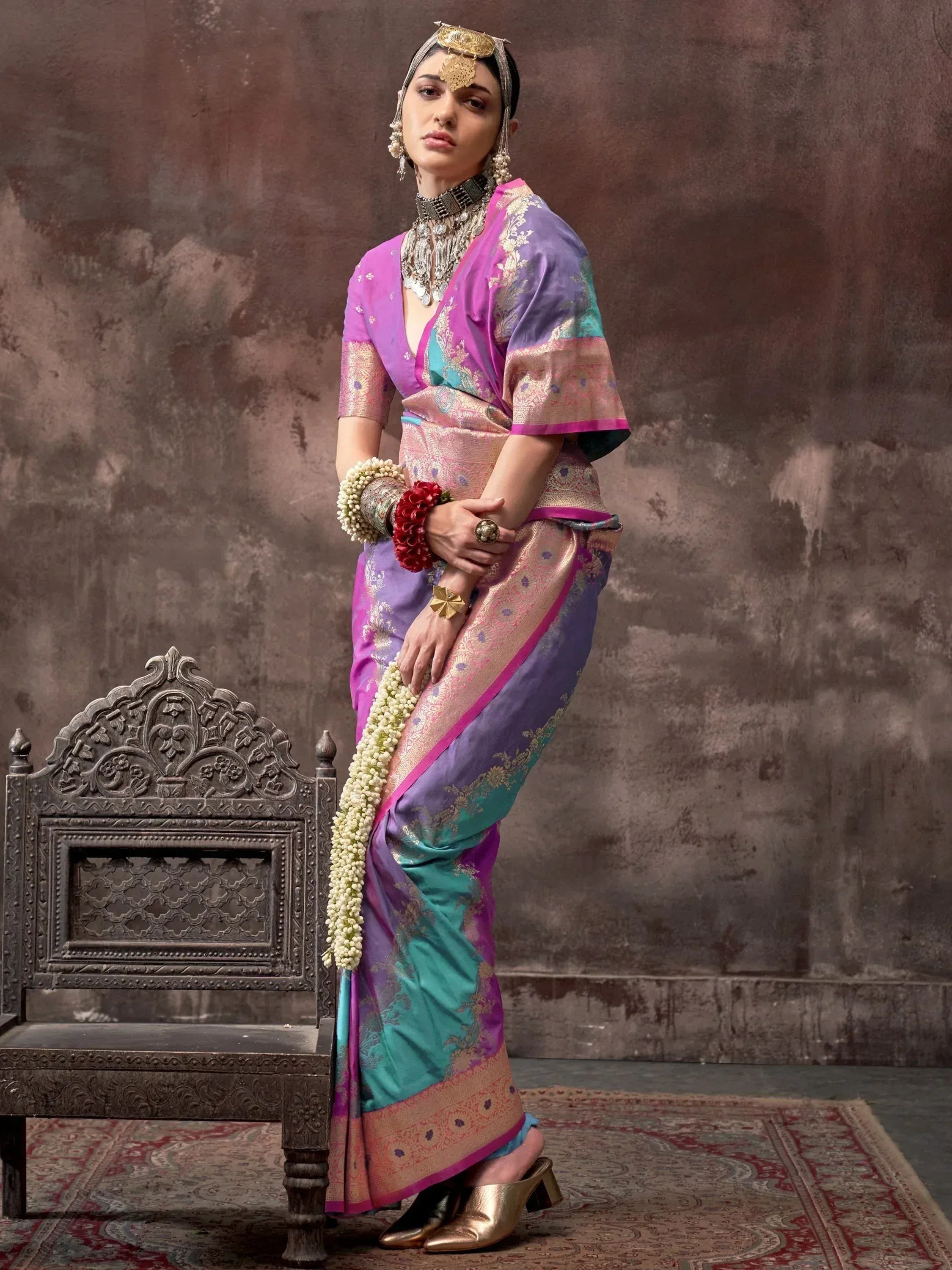 Woman in vibrant purple and teal designer saree with gold embroidery, traditional jewelry, and floral garland