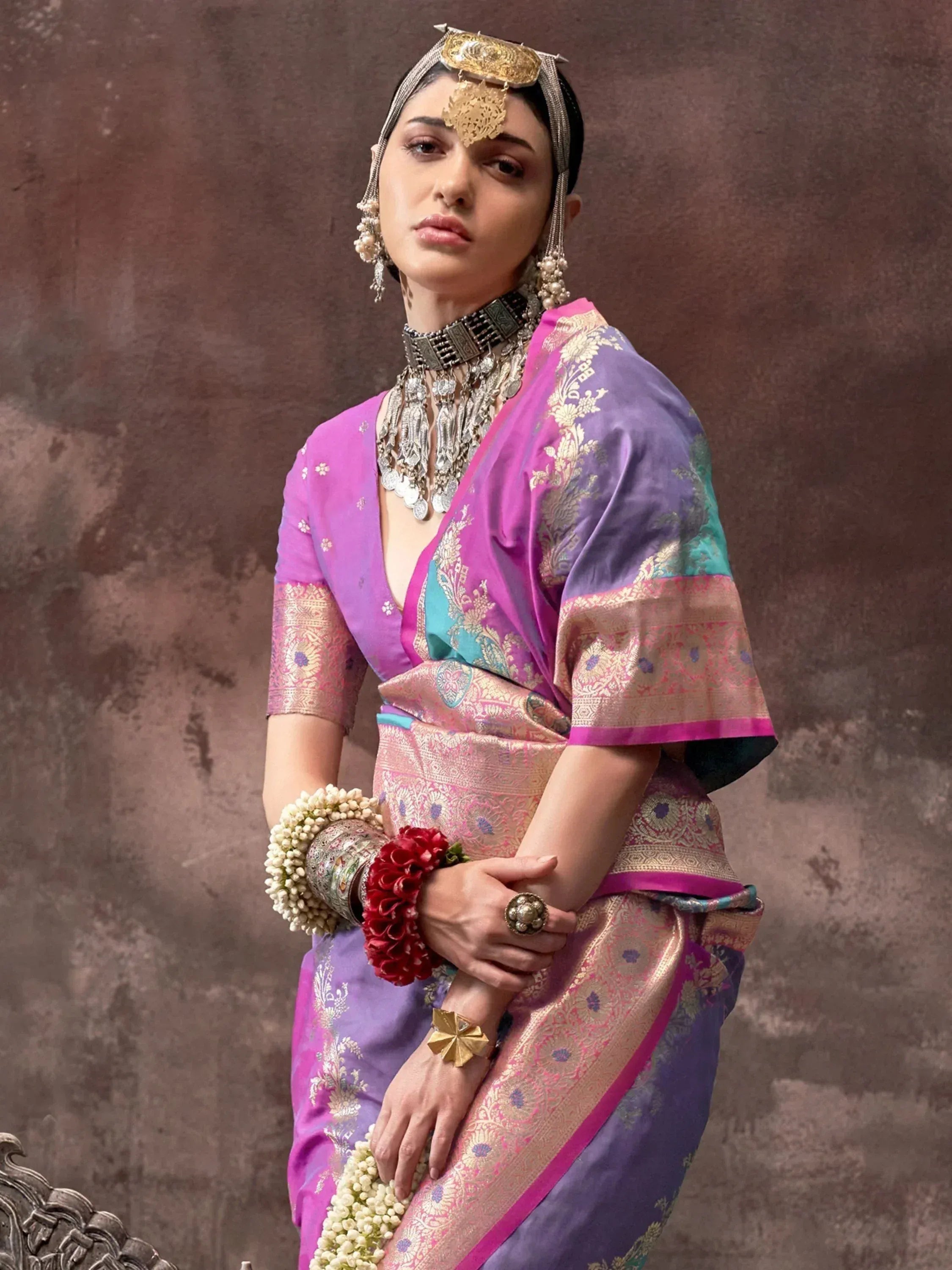 Woman in purple and pink designer saree with gold embroidery, traditional jewelry, and floral accessories