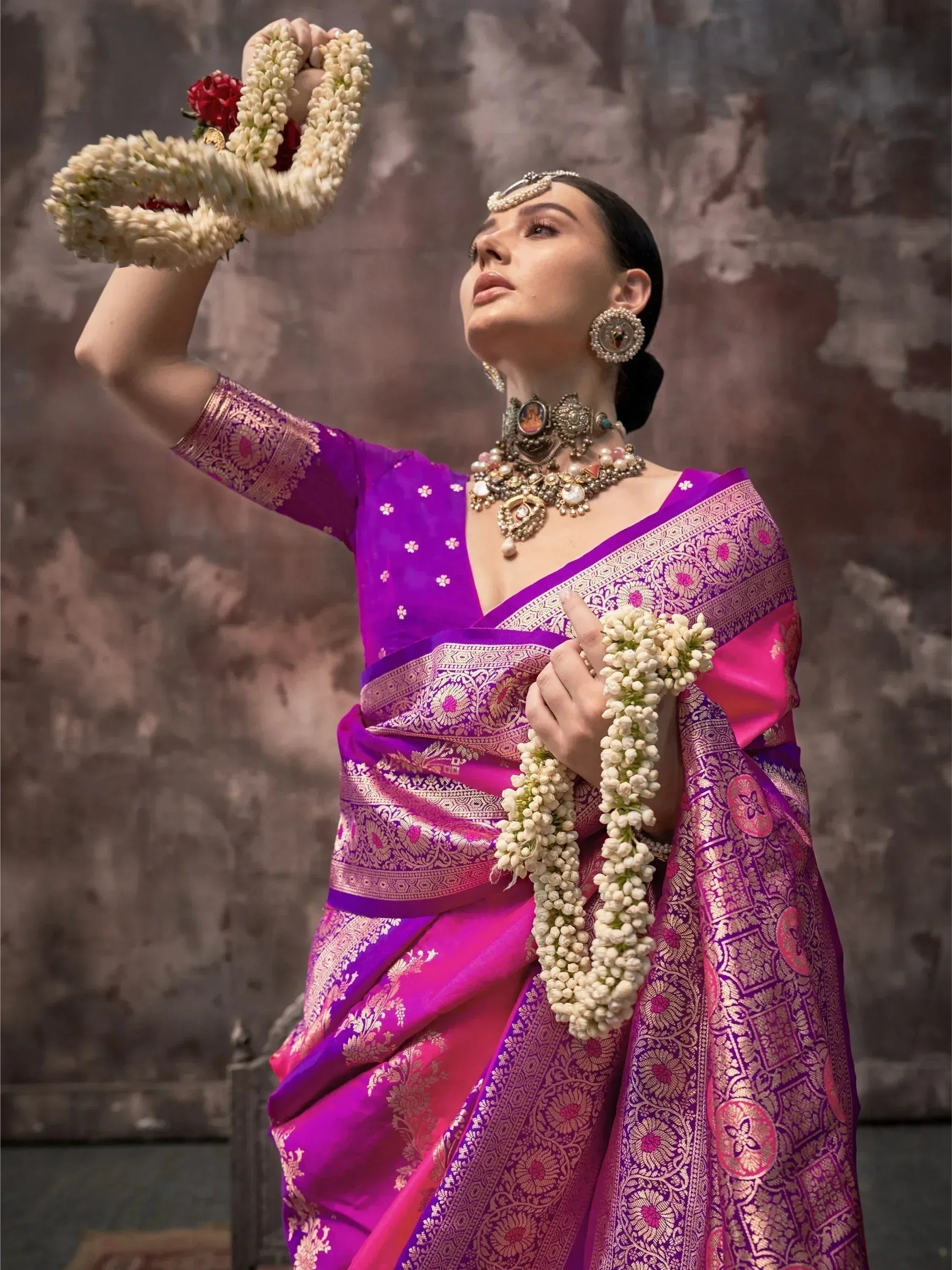 Woman in vibrant purple and pink designer saree with gold embroidery, holding floral garlands, styled with traditional Indian jewelry.