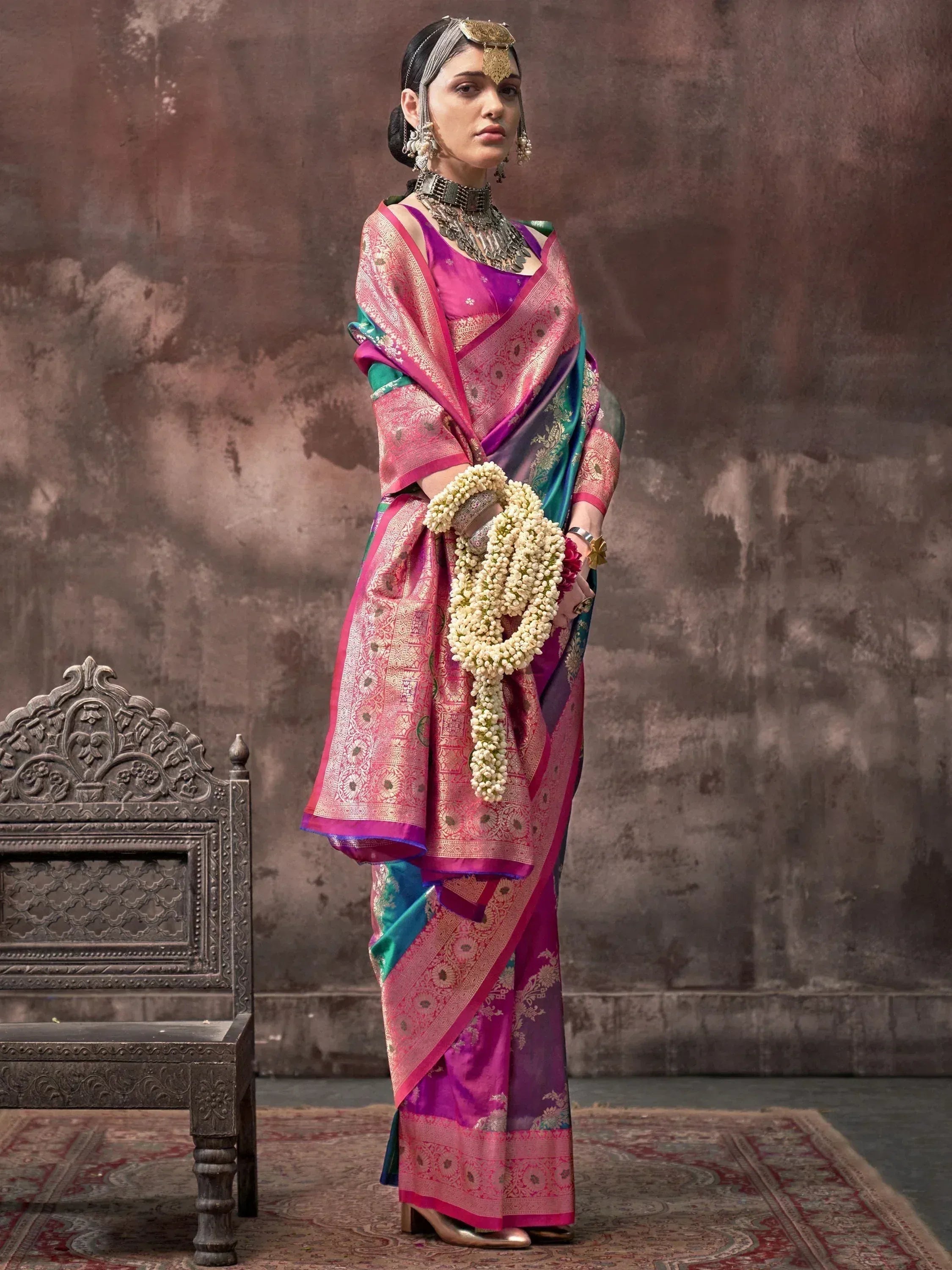 Woman in vibrant pink and purple Banarasi saree with gold embroidery, traditional jewelry, and floral garland