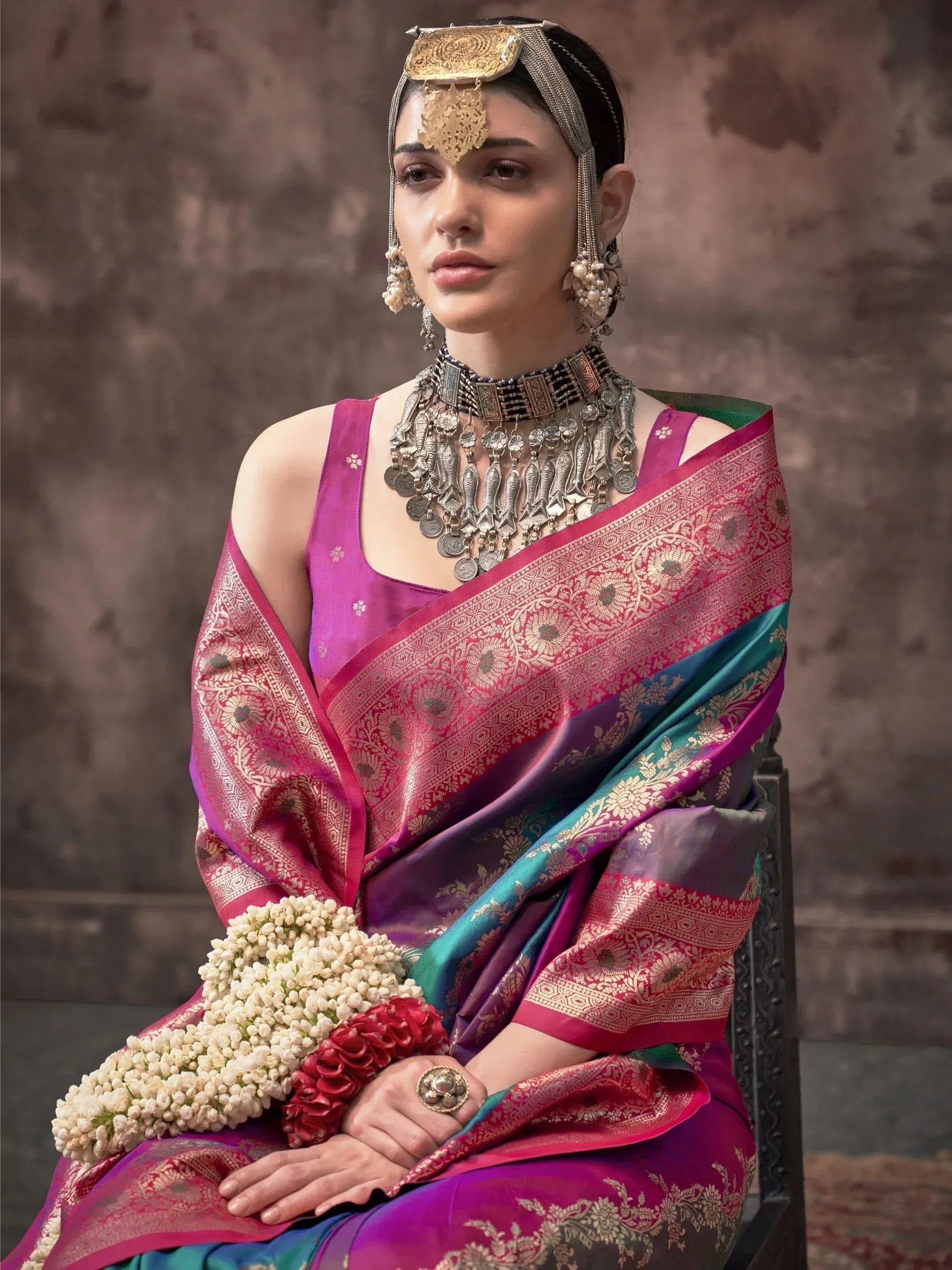 Woman in vibrant pink and teal silk saree with gold embroidery, traditional Indian jewelry, and floral garland.