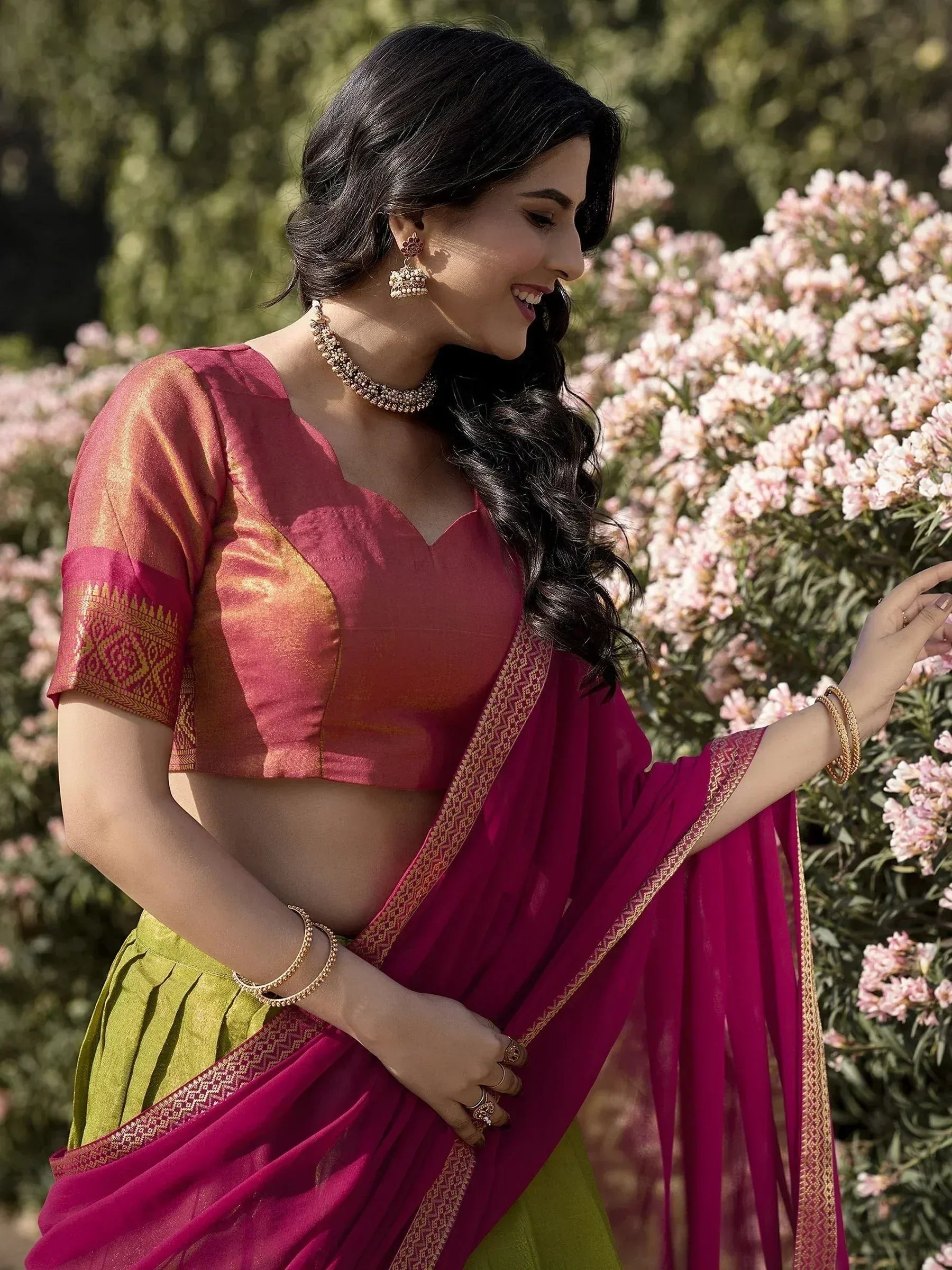 Woman in vibrant pink and green ethnic saree with gold border, styled with traditional jewelry in garden setting