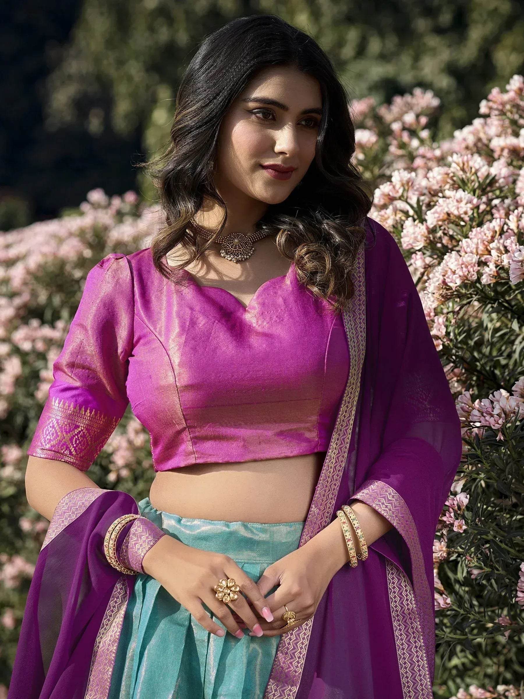 Purple and teal Indian ethnic lehenga choli with gold embroidery, worn with dupatta and jewelry in garden setting