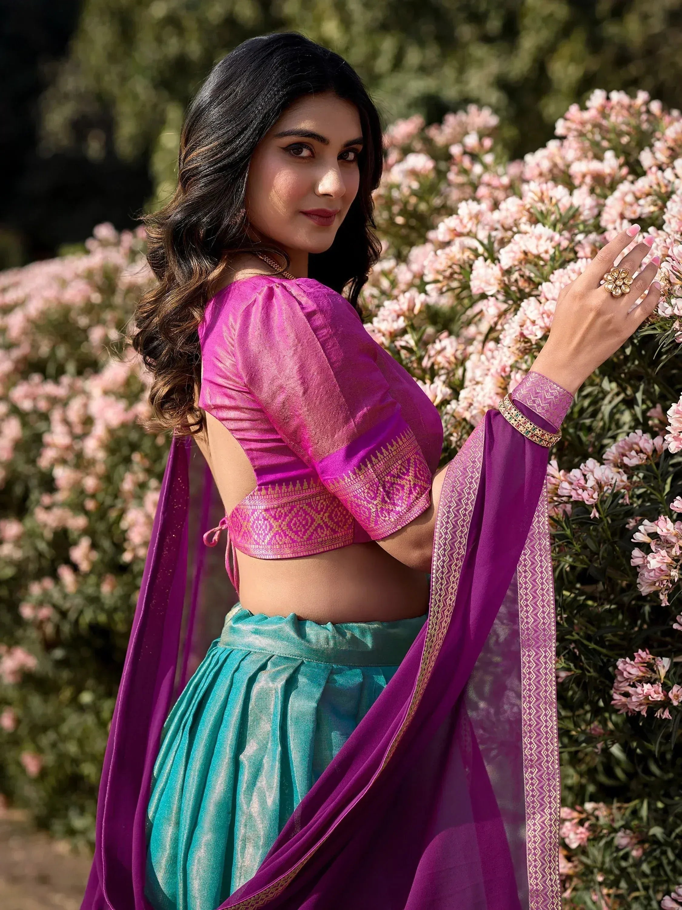 Woman in vibrant pink and teal ethnic lehenga choli with gold embroidery, posing near pink flowers