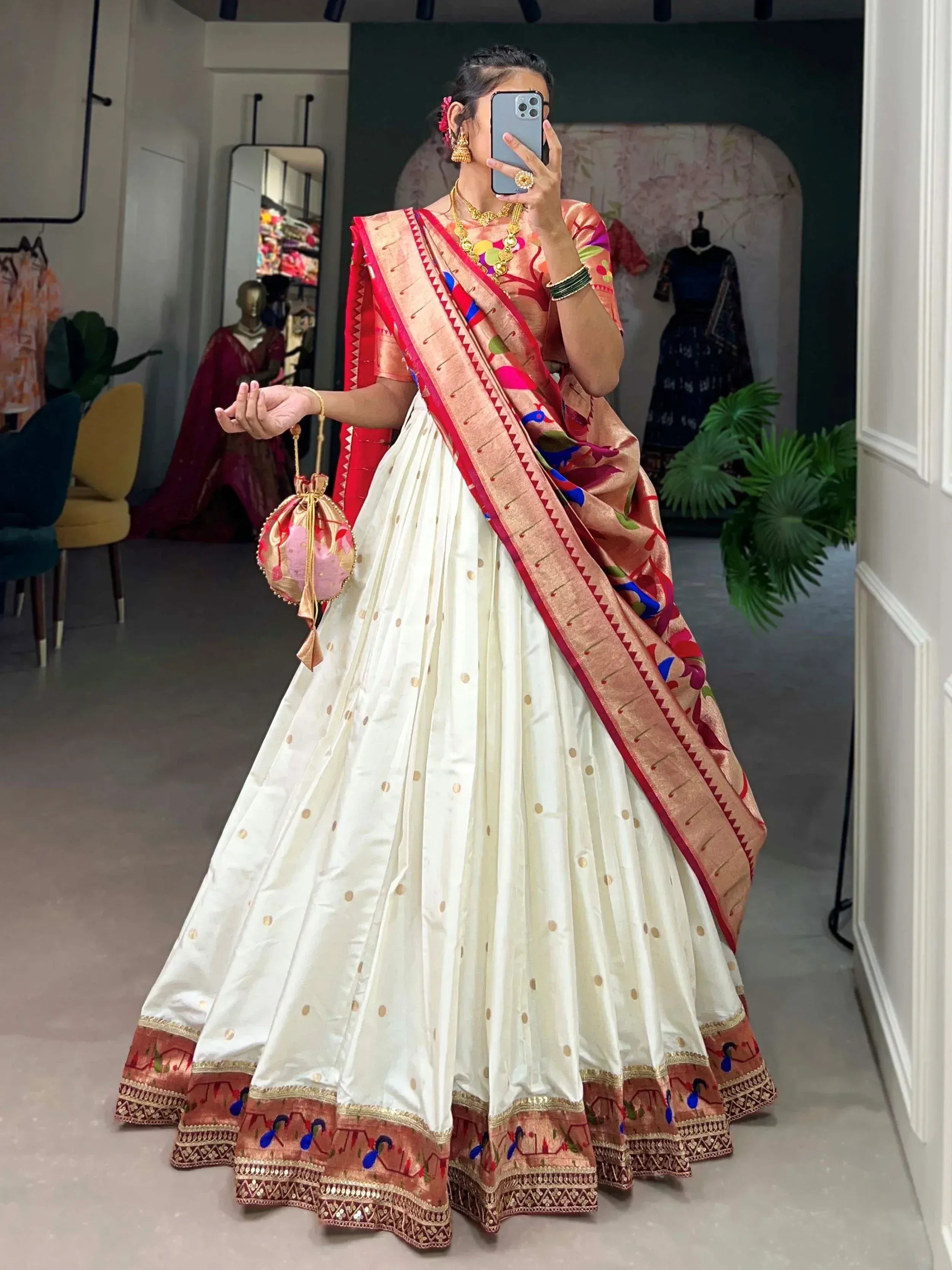 Woman in white lehenga with colorful floral dupatta, gold jewelry, and potli bag, taking mirror selfie in boutique