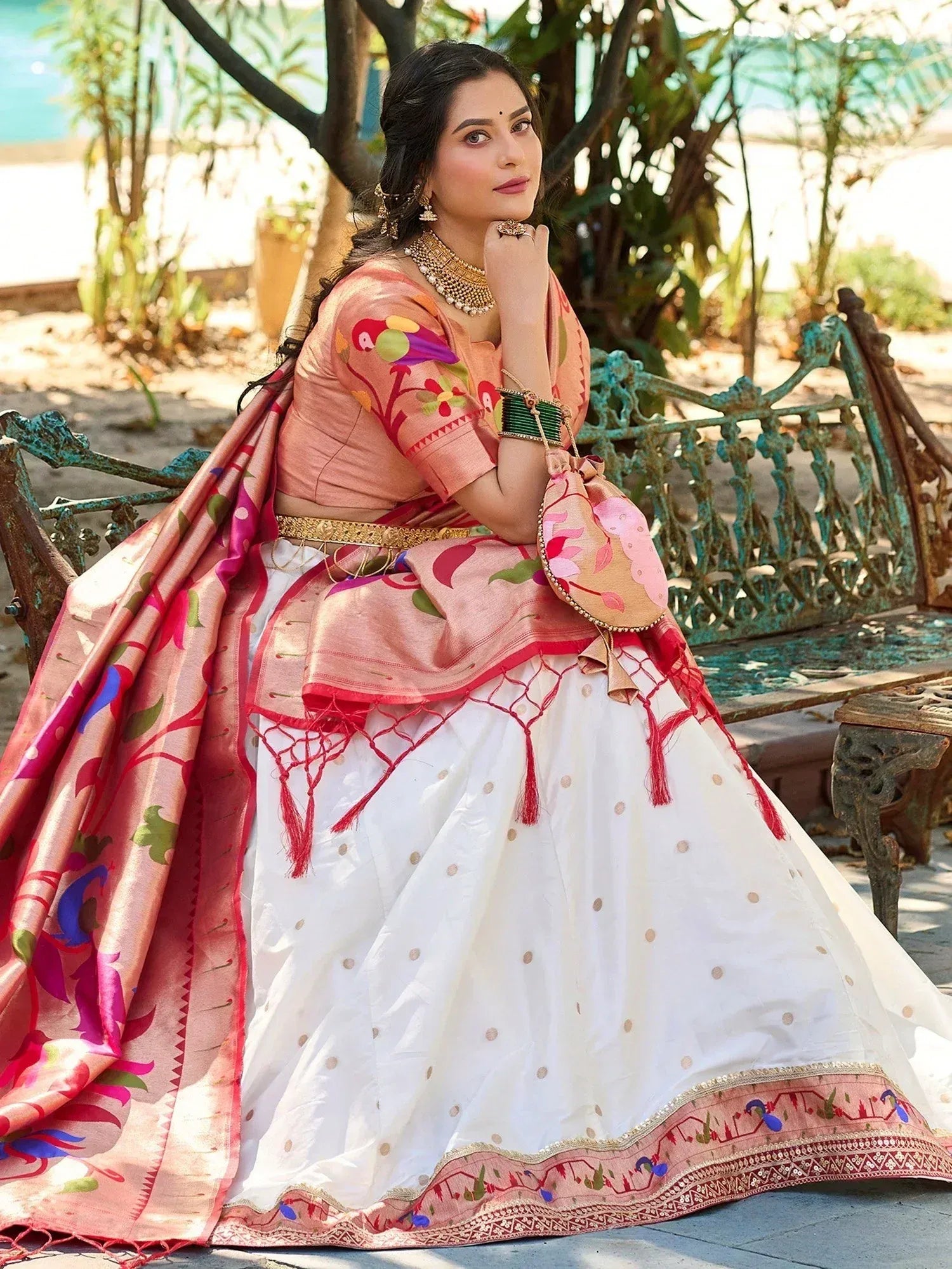 Woman in pink and white embroidered lehenga choli with floral dupatta, sitting on garden bench