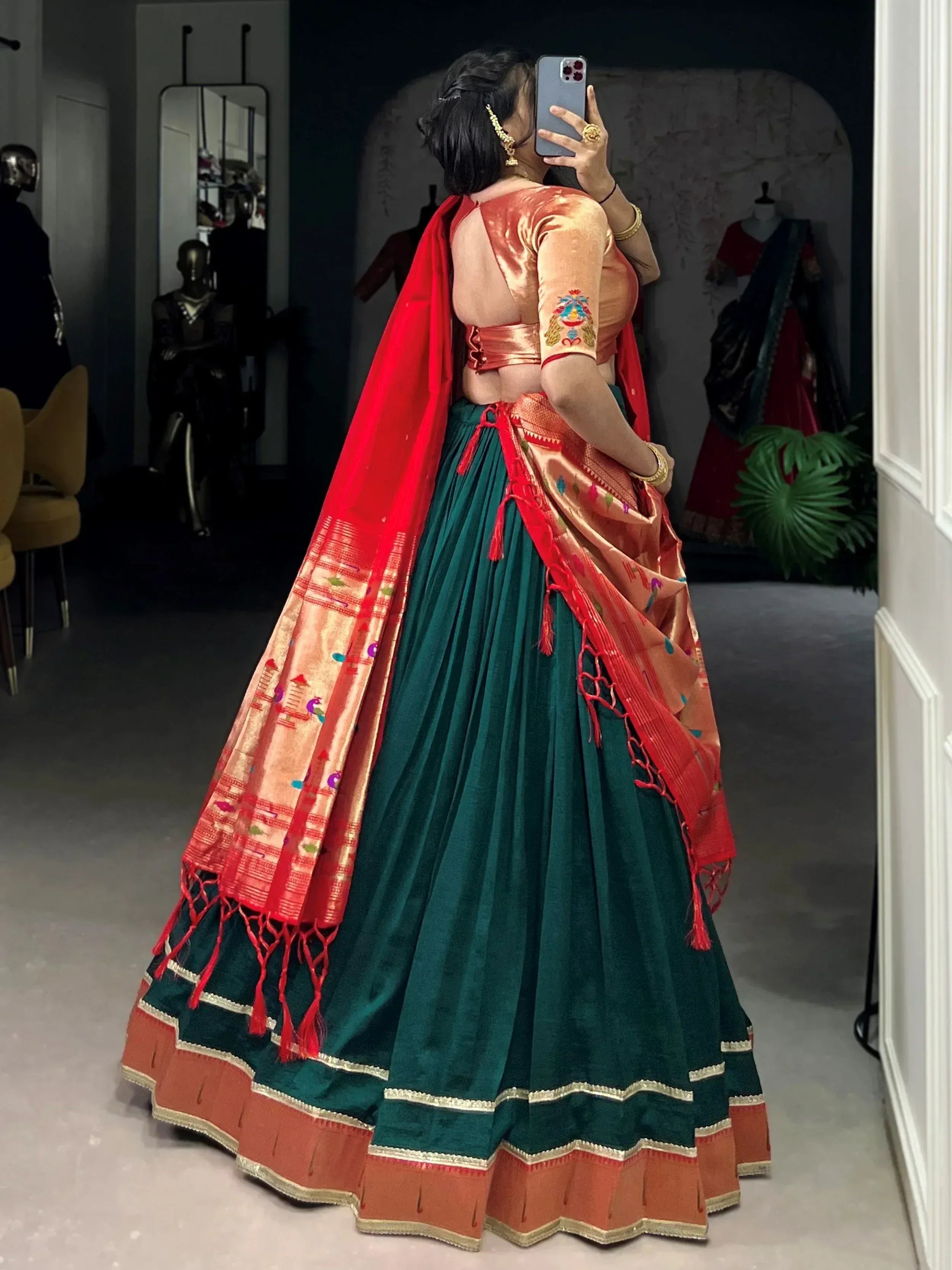 Woman in green velvet lehenga with peach blouse and red dupatta, taking mirror selfie in boutique