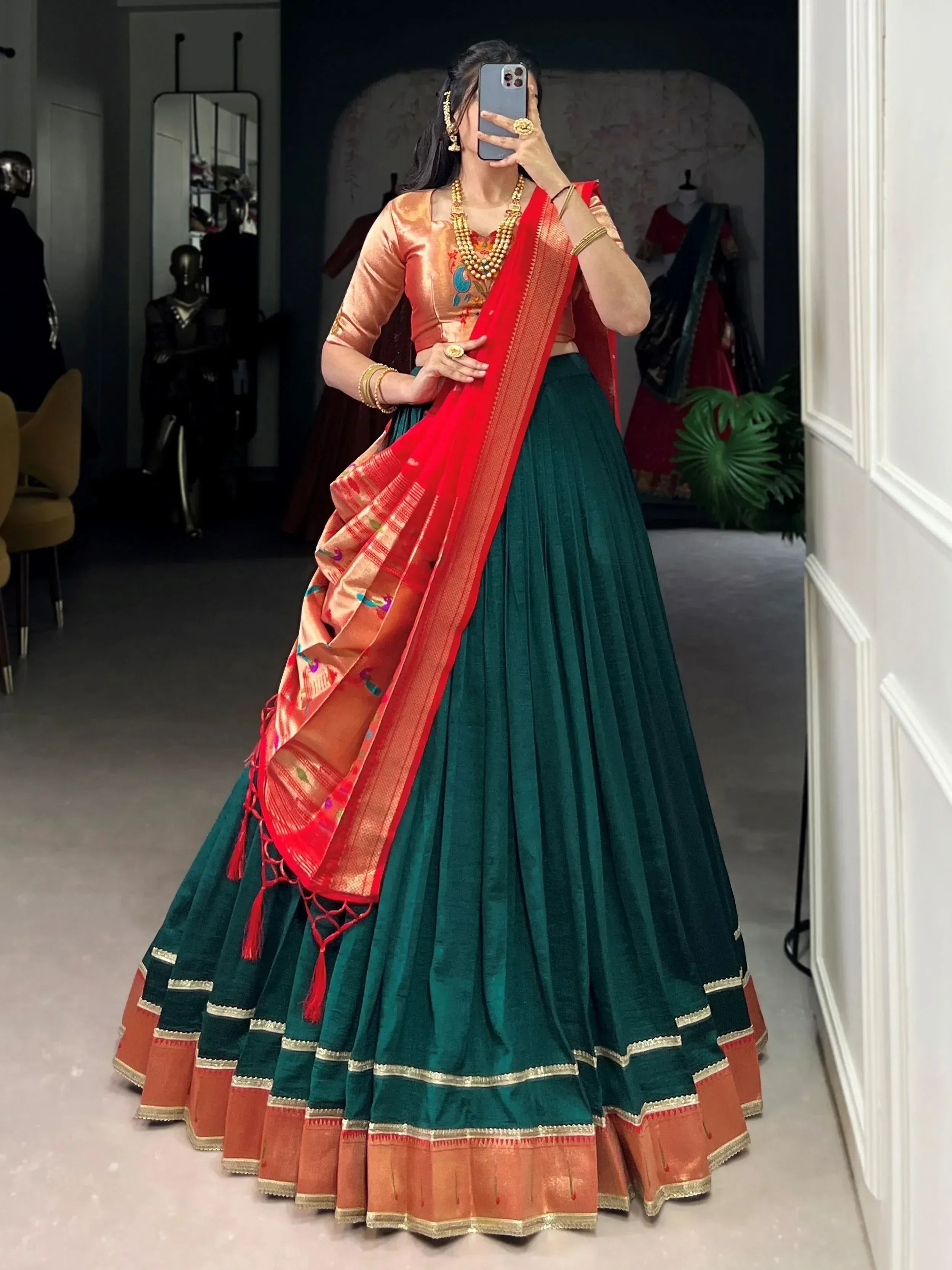 Woman in green velvet lehenga with red dupatta, peach blouse, gold jewelry, taking mirror selfie
