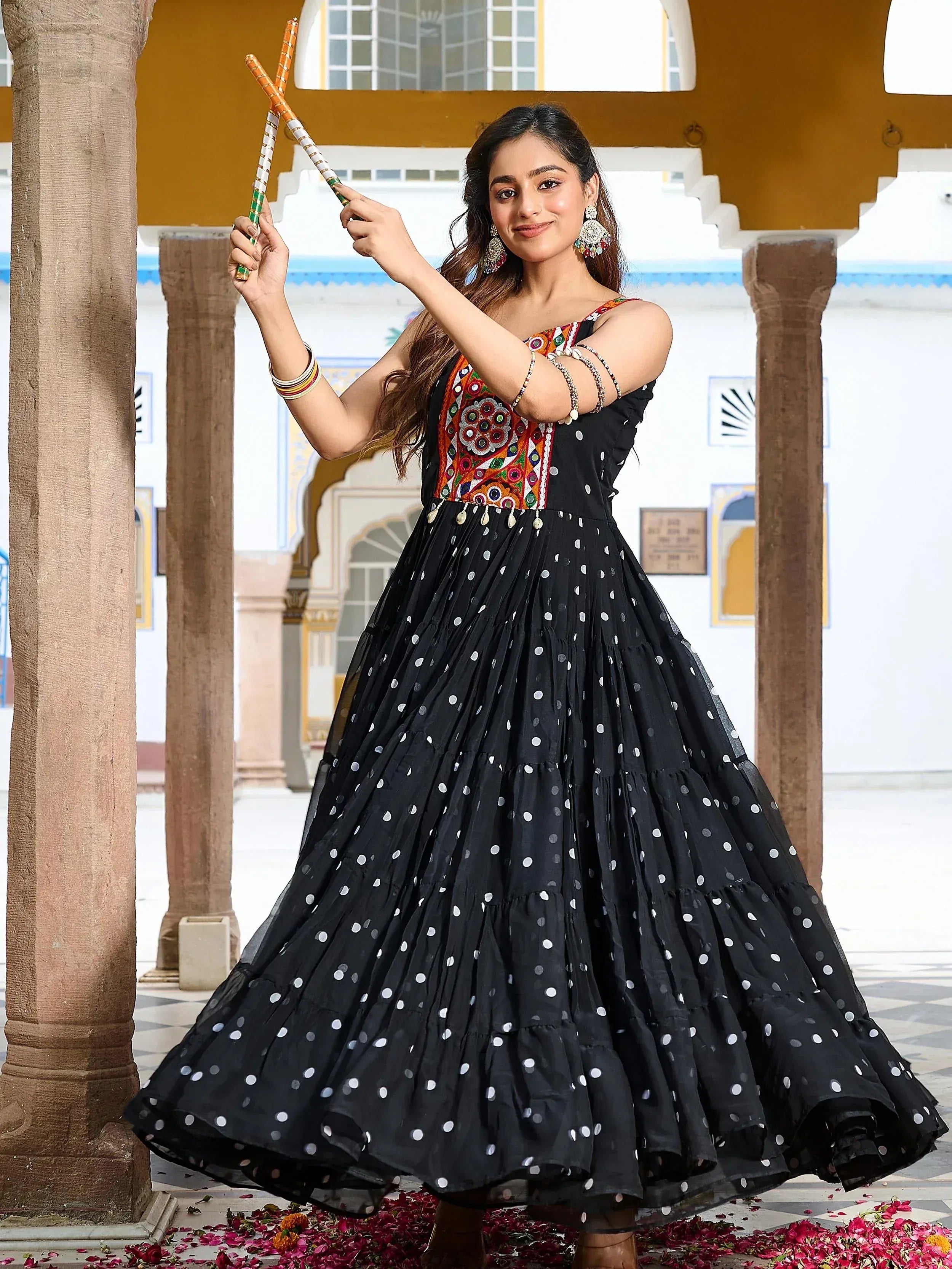 Woman in black polka dot ethnic gown with colorful embroidery, holding decorative sticks, traditional Indian attire
