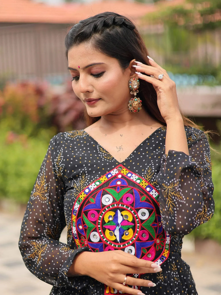 Woman in black ethnic gown with colorful mirror work, styled with traditional earrings and braided hair.