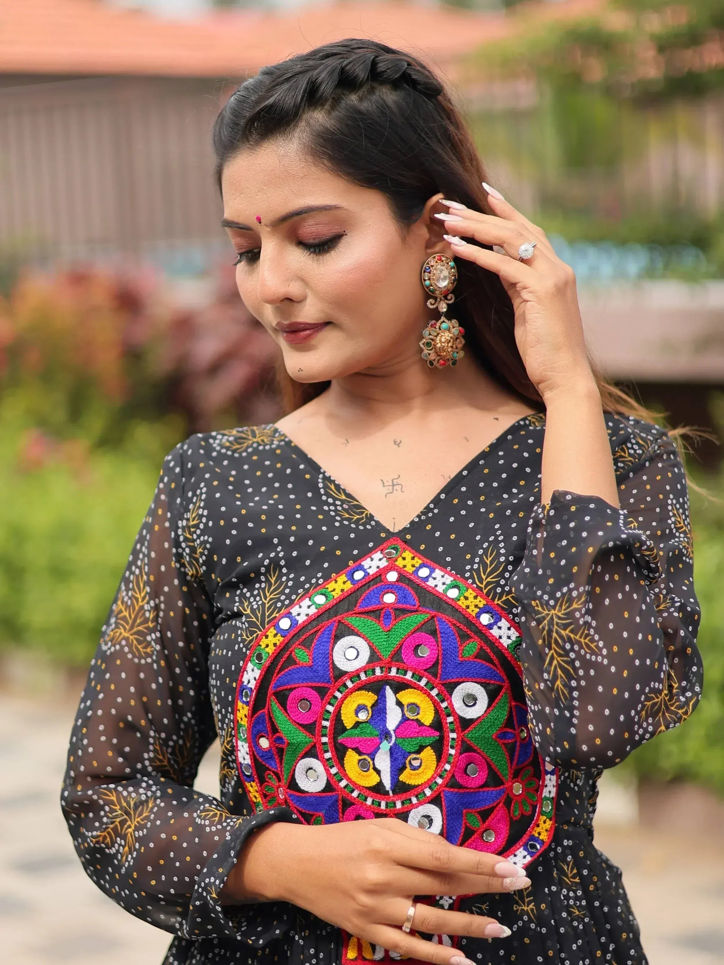 Woman in black ethnic gown with colorful mirror work, styled with traditional earrings and braided hair.