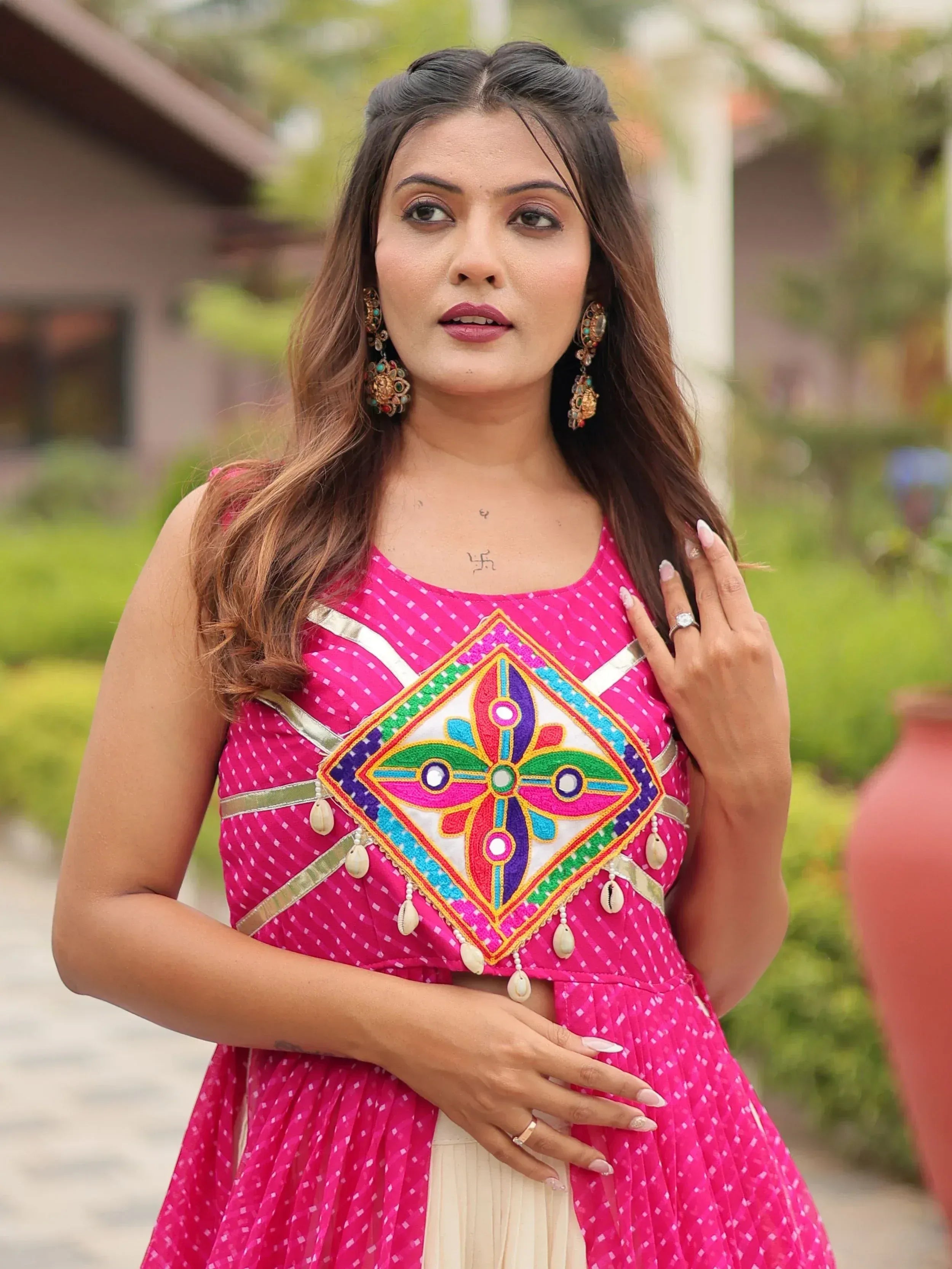 Woman in vibrant pink ethnic gown with colorful mirror embroidery and shell tassels, perfect for Indian celebrations.