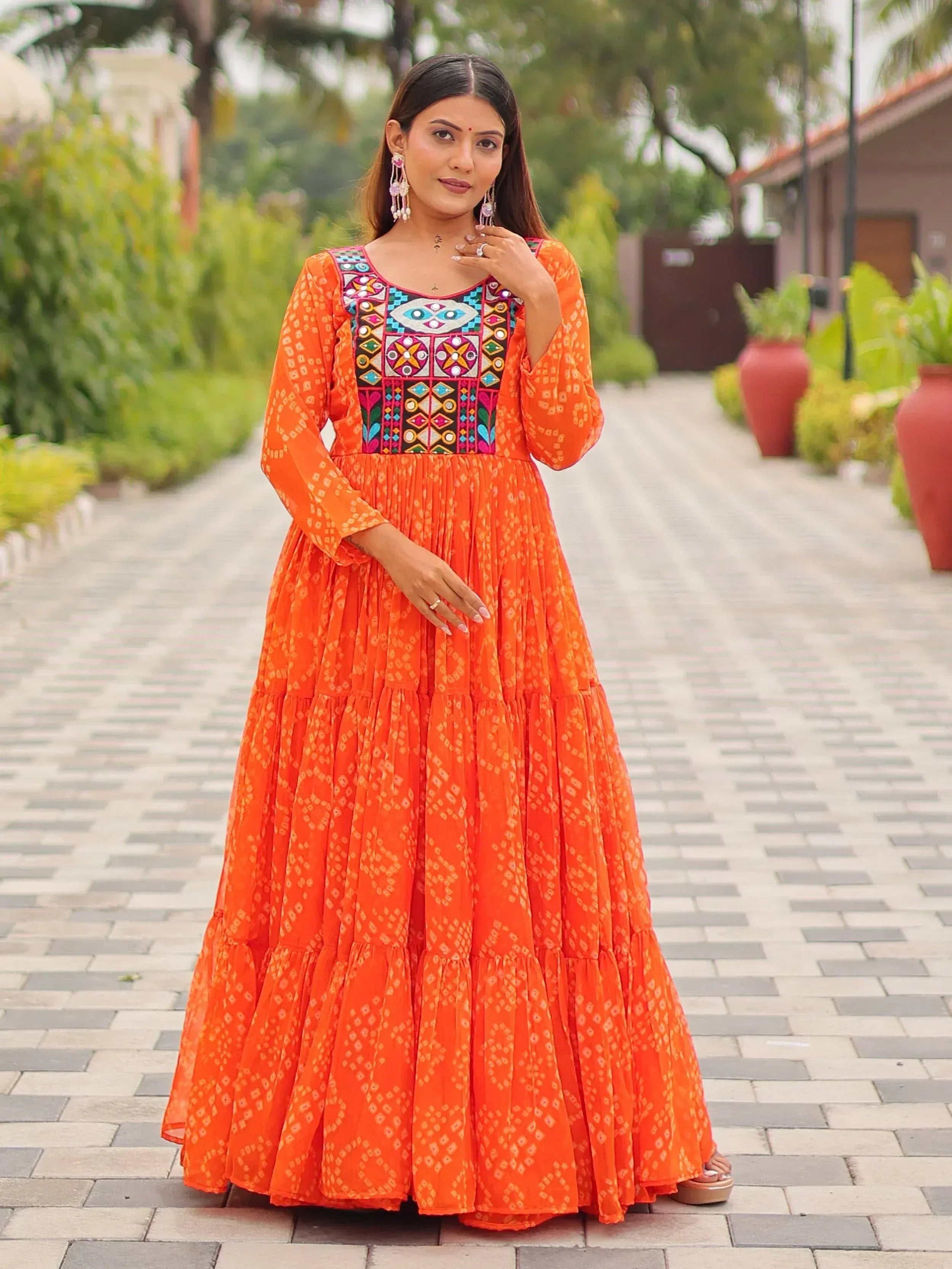 Vibrant orange ethnic gown with colorful mirror work and tiered skirt, perfect for Indian festivities