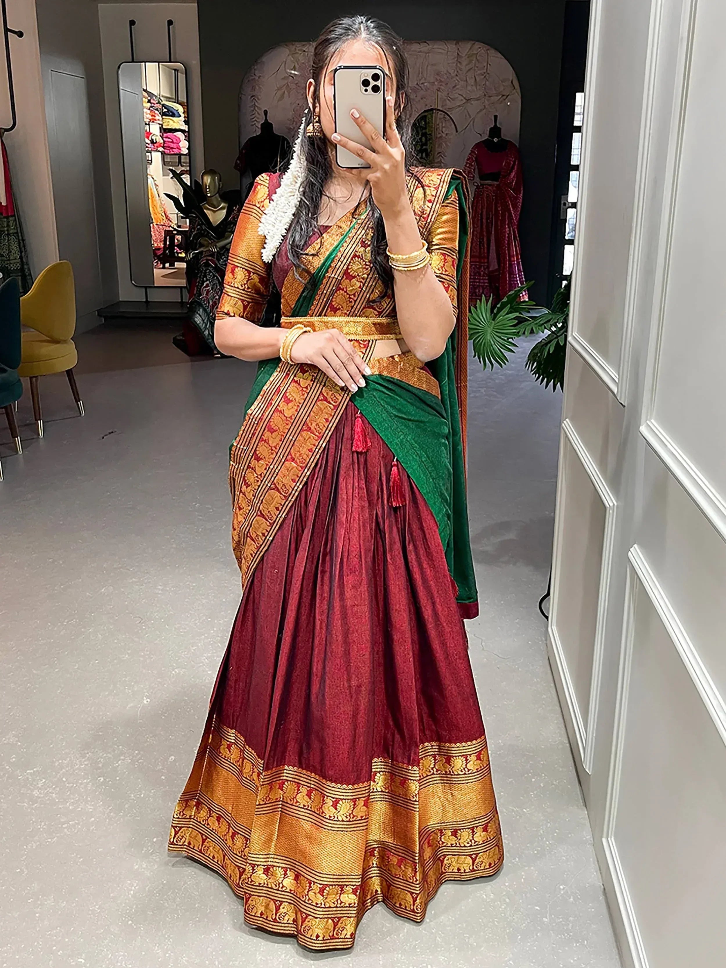 Woman in maroon and green designer lehenga choli with gold embroidery, taking mirror selfie in boutique